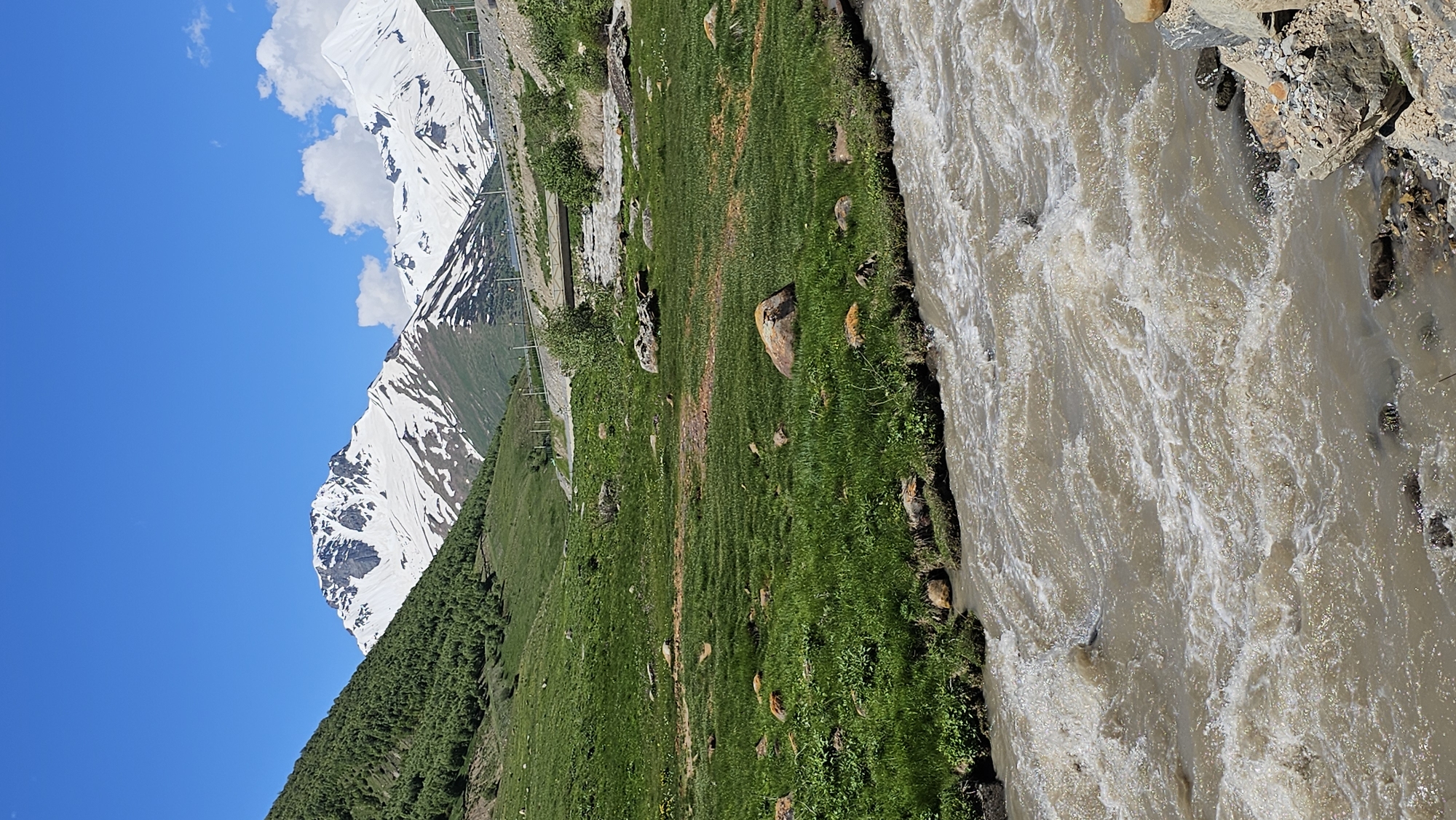 A vertical, eye-level shot captures a rushing, muddy river in the foreground, its turbulent waters filled with white foam and rapids. The river flows from the bottom right toward the bottom left of the frame. Scattered rocks line the immediate banks of the river, some submerged in the water.
Beyond the river, a lush green grassy meadow stretches uphill, dotted with various sized rocks. The meadow is bordered by a steep, tree-covered slope on the left, leading up to snow-capped mountains in the background. The peaks of these mountains are jagged and rugged, with patches of snow clinging to their upper reaches. A clear, bright blue sky with a few scattered white clouds dominates the upper portion of the image.
In the middle ground, a road curves through the landscape, with power lines and poles visible alongside it. A small concrete bridge spans a section of the landscape behind the road. The scene depicts a natural mountain landscape, likely in a rural or remote area, given the lack of prominent man-made structures beyond the road and bridge. The location is identified as Kamskho, Russia.