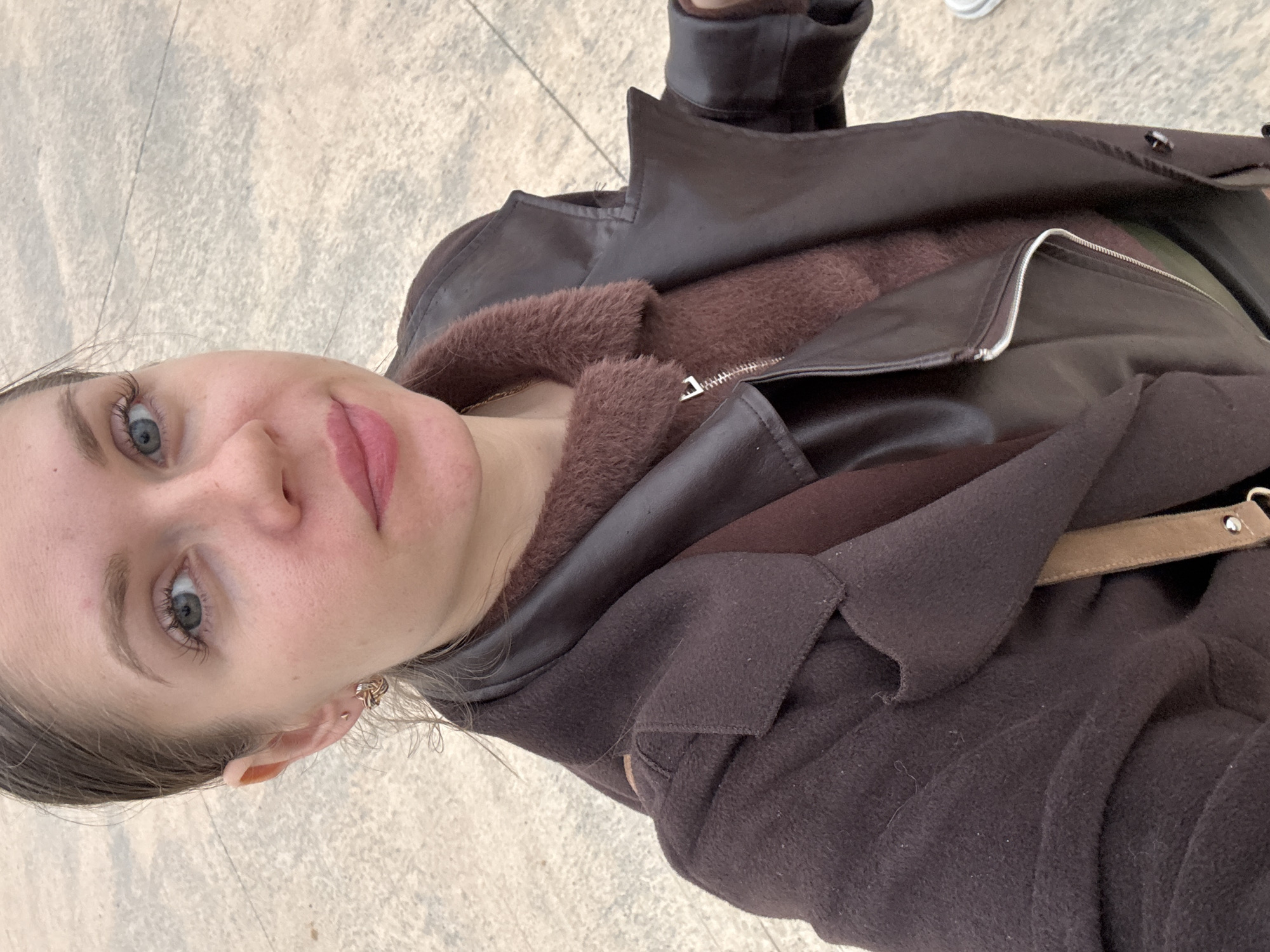 A fair-skinned woman with light-colored eyes is the primary subject of this selfie-style photograph. She has a serene expression with her lips slightly pursed. Her hair is light brown and pulled back. She is wearing a brown, multi-layered jacket with faux fur trim, which partially obscures the background.
The background shows a paved surface, indicative of a sidewalk or public space. There is minimal background detail visible to make a determination about the location in the city of Batumi, Georgia.