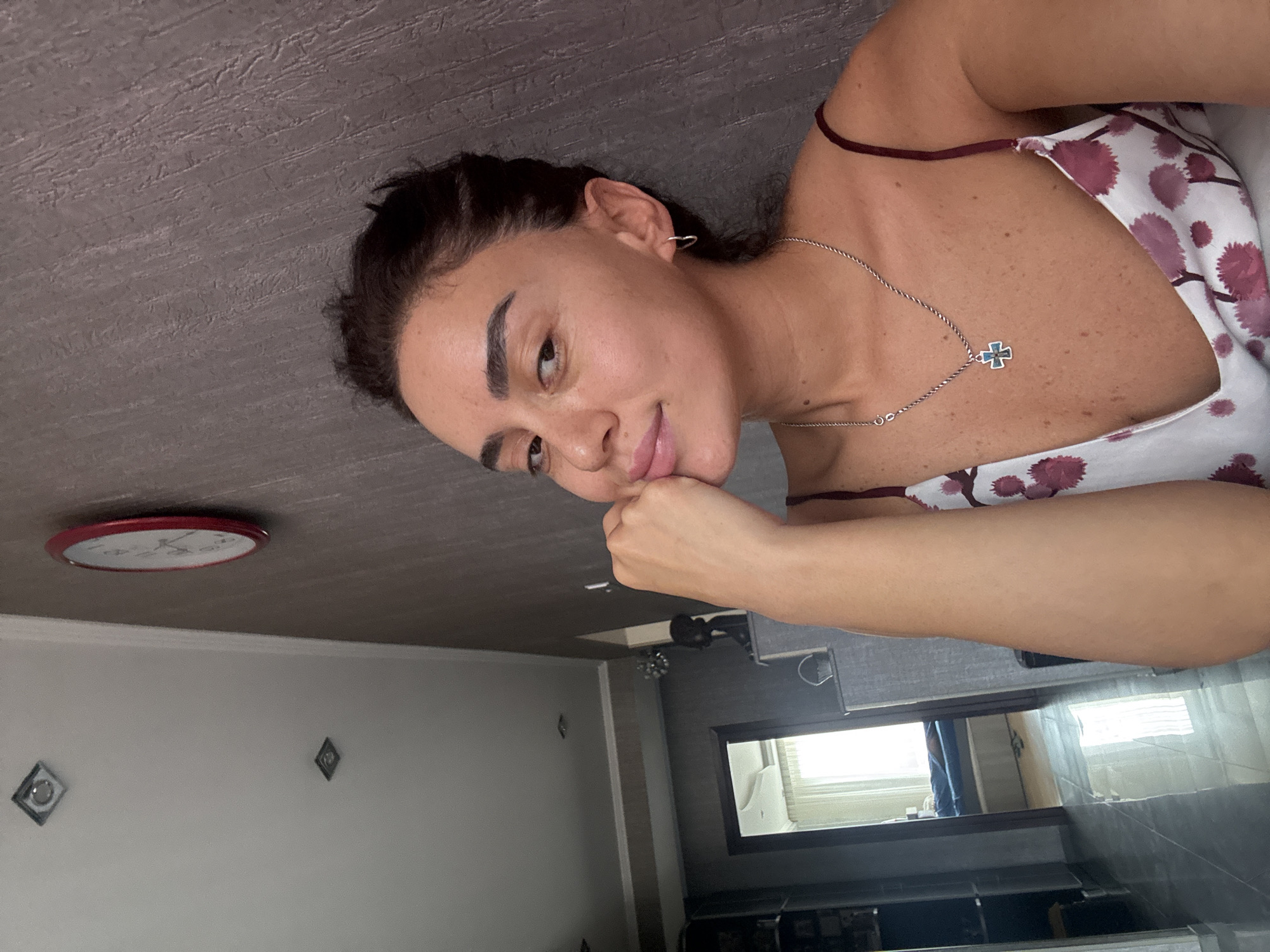 A medium shot shows a woman with dark hair pulled back, resting her chin on her hand. She is wearing a white camisole with a floral pattern and a silver necklace with a cross pendant. Her left ear has a small hoop earring. The background is a textured grey wall. To the left of the woman, a doorway leads to another room where a bed with blue bedding is visible. The ceiling has recessed lighting fixtures, and a round red clock hangs on the wall. The scene appears to be an indoor domestic setting, possibly a bedroom or living area, in Tbilisi, Georgia.