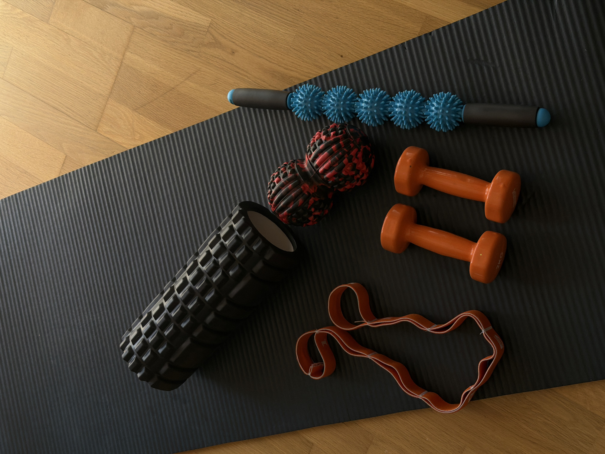An overhead, slightly angled shot captures a collection of fitness and recovery equipment arranged on a black yoga mat, which lies partly on a light brown wooden herringbone floor. The setting appears to be indoors, potentially a home workout space in Budva, Montenegro.
On the left side of the mat, a black foam roller with a prominent grid pattern is positioned. Towards the center, a red and black mottled double massage ball is visible. Above it, a blue spiky massage roller stick with black handles extends horizontally across the upper part of the mat.
To the right, two orange dumbbells are neatly placed, one above the other. The upper dumbbell clearly displays "2KG" printed on its side, indicating its weight. Below the dumbbells, an orange resistance band is laid out, forming a loose, undulating shape.
The ridged texture of the black yoga mat is clearly visible, contrasting with the smooth, polished surface of the wooden floor. The overall arrangement suggests a space prepared for, or recently used for, a workout, stretching, or physical therapy session. The lighting is even, suggesting a bright daytime environment, though no specific time of day or weather conditions can be definitively determined. No people are present in the image, and thus, no emotions are conveyed.