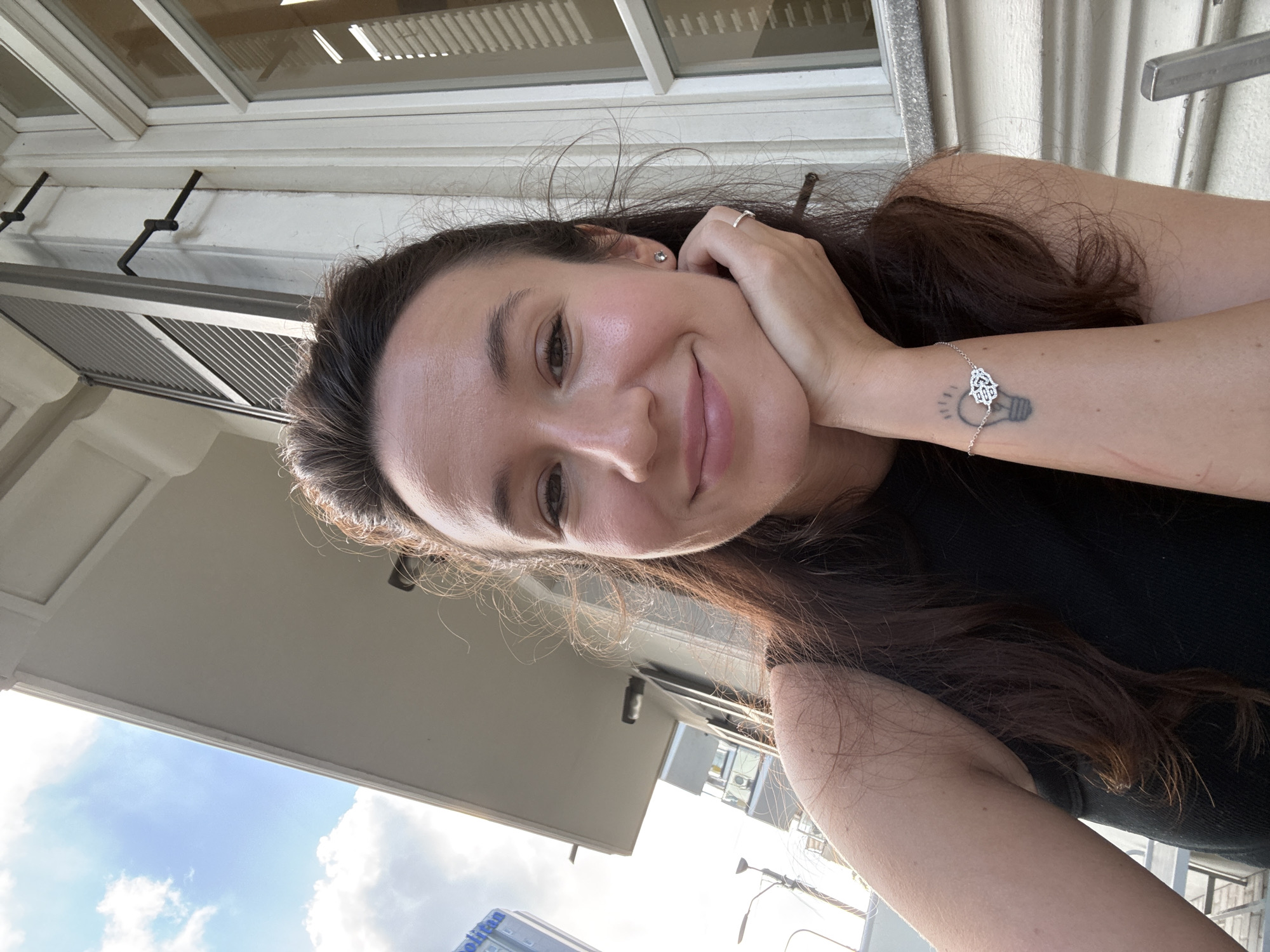 A woman with long, dark hair and a black top is captured in a selfie from the chest up. She rests her chin on her hand, her left arm adorned with a bracelet and a lightbulb tattoo on her forearm. Her expression is a relaxed smile, and she's wearing a silver ring on her ring finger and a small earring on her left ear. Behind her, a building with white shutters and a gray awning is visible, suggesting an outdoor setting, possibly a cafe or balcony. The sky above is blue with scattered clouds, indicating daytime. The building's architecture, particularly the shutters and awning, is consistent with structures found in Tel-Aviv, Israel, a city known for its Bauhaus and international style architecture, where such outdoor seating areas are common.