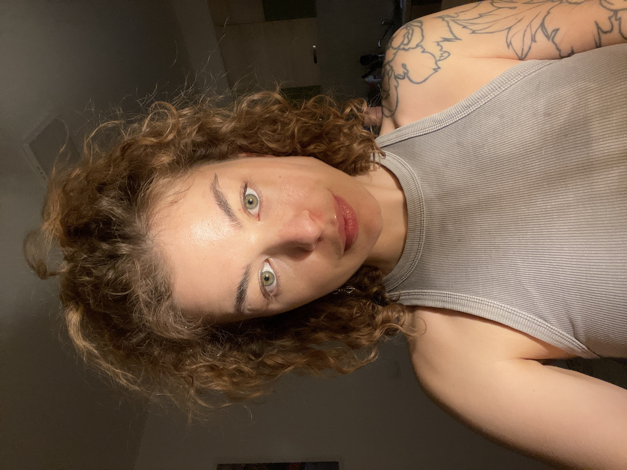 A woman with curly red hair and light green eyes, wearing a gray ribbed tank top, is angled towards the camera. She has a tattoo on her upper left arm and shoulder. The background is dimly lit, revealing glimpses of a door and an unidentified object. No specific location is discernible. No specific actions or interactions are occurring. The environment appears to be indoors.