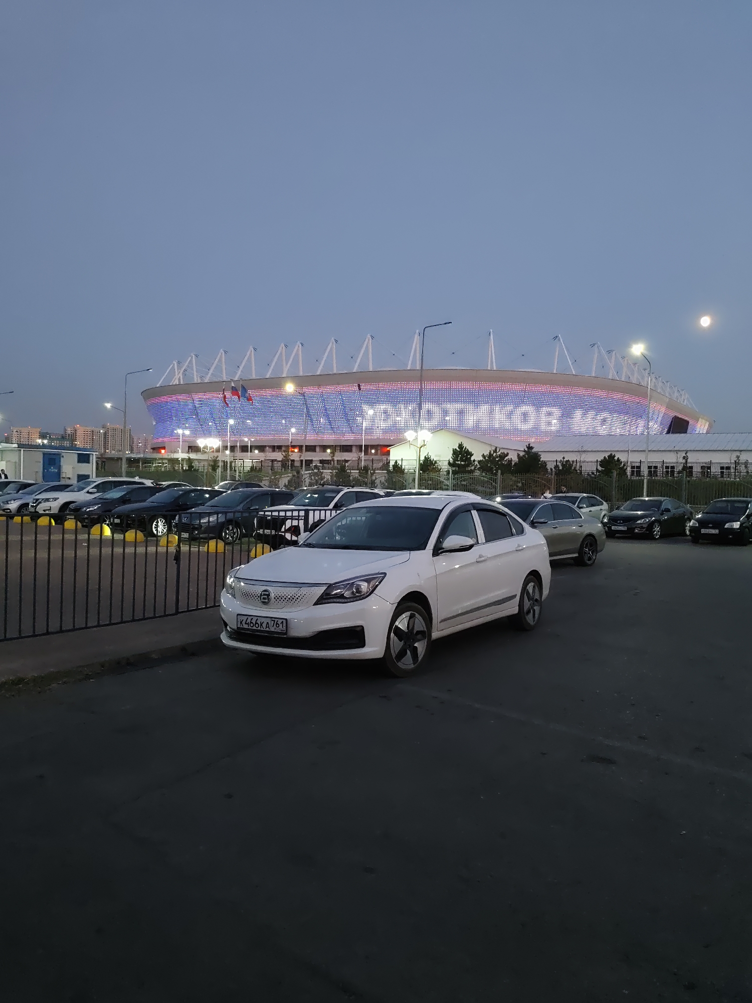 A nighttime, eye-level, medium shot captures a parking lot in front of a brightly lit stadium in Rostov-on-Don, Russia. The stadium, adorned with an illuminated pattern of blue and red lights, displays Russian text that appears to read "РОСГОССТРАХ" (ROSGOSTRAKH) and "РОСТОВ-НА-ДОНУ" (ROSTOV-ON-DON). Two Russian flags are visible on the stadium's facade.
In the foreground, a white electric sedan, identified by its license plate K466KA 761, is parked. Behind it, a line of various sedans and SUVs are parked in a lot bordered by a black metal fence. Trees and low-rise buildings are visible beyond the fence, with the stadium serving as the dominant backdrop. The sky is a muted, dusky blue, with a full moon present on the right side of the frame. There are no people clearly visible in the image. The overall scene suggests a quiet evening after an event, with the stadium's lights still active.