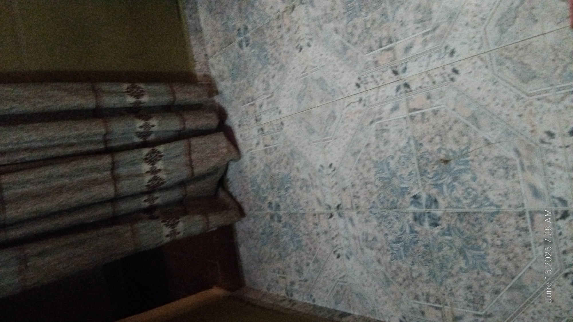 The image shows a tiled floor with a patterned design. A curtain is visible in the upper portion of the image, partially obscuring a dark space. The lighting is dim, and the overall scene appears to be indoors. There are no people or discernible actions taking place. The timestamp "June 15, 2026 7:28 AM" is present in the lower left corner.