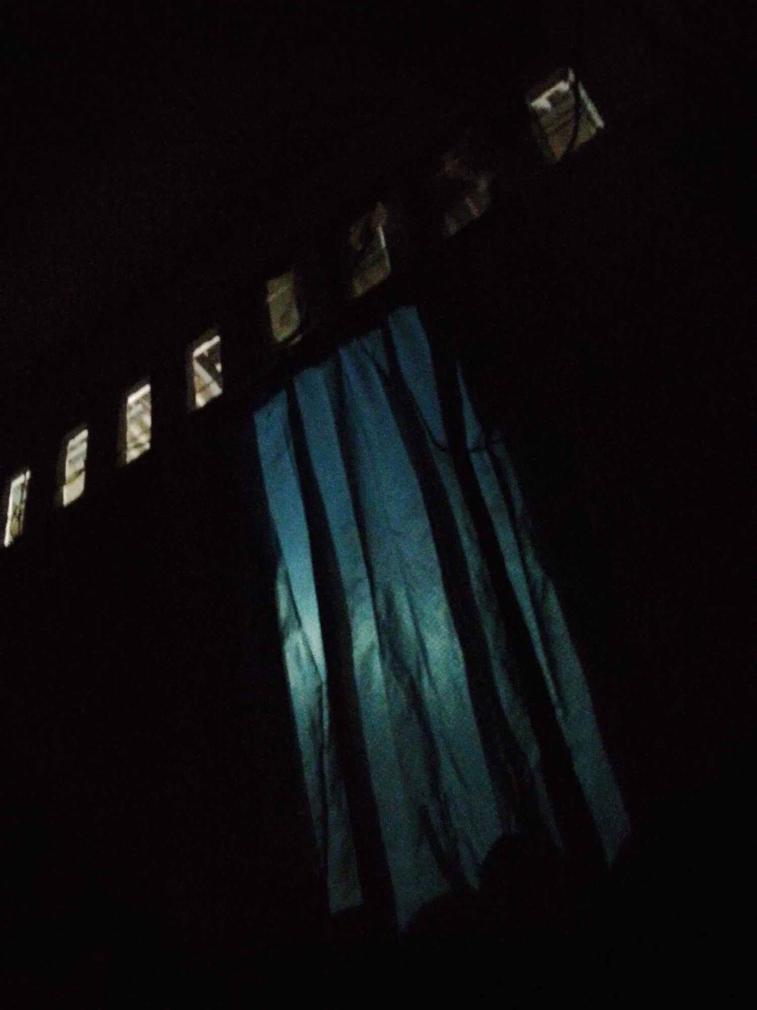 This is a low-light indoor scene, possibly a room in Bukit Ketok, Indonesia. A curtain with vertical stripes, predominantly blue-green in color, hangs beneath a series of small, rectangular openings in the upper part of the wall. Light is visible through these openings, which may be windows or vents. The remainder of the image is dominated by dark, undefined space. There are no people visible.