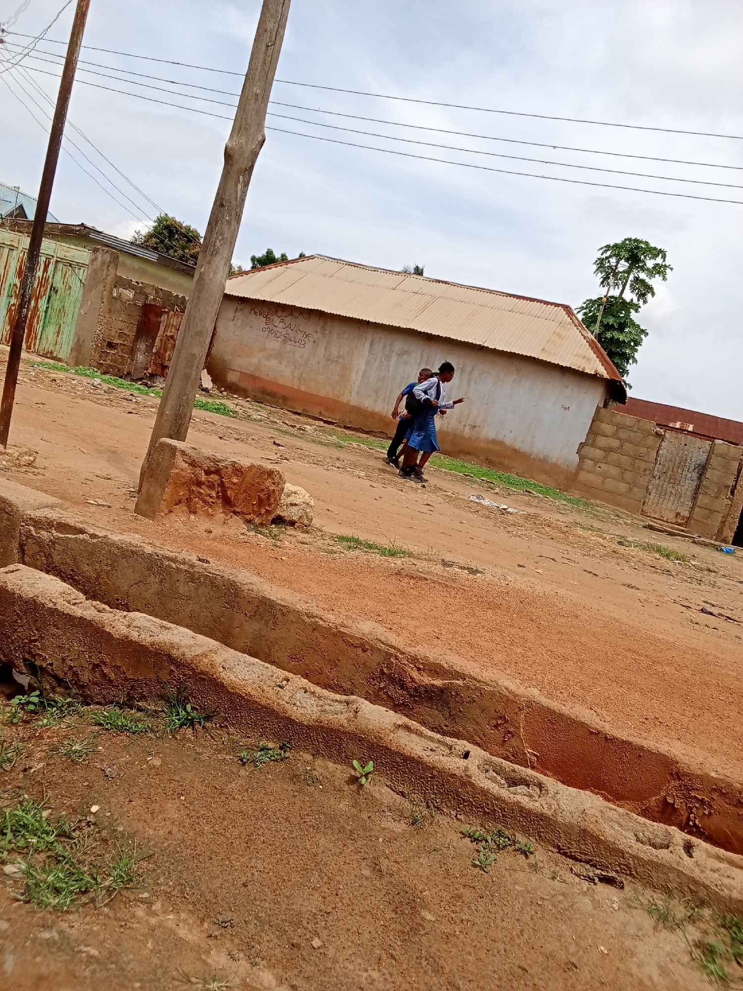 The image depicts a street scene in Narayi, Nigeria. In the mid-ground, two individuals, appearing to be schoolchildren, are walking away from the camera. The individual in the foreground is wearing a blue skirt and a white shirt, carrying a backpack. The other individual, partially obscured, is wearing dark clothing and also appears to be carrying a backpack. They are walking on a dirt path in front of a white-walled building with a corrugated metal roof. To the left of the building is a green metal gate with visible rust. A wooden utility pole stands prominently in the left mid-ground. In the foreground, there are concrete structures that appear to be drainage channels, filled with dirt and sparse vegetation. The background shows more buildings constructed with brick and corrugated metal roofing, as well as overhead power lines and a tree. The sky is overcast.