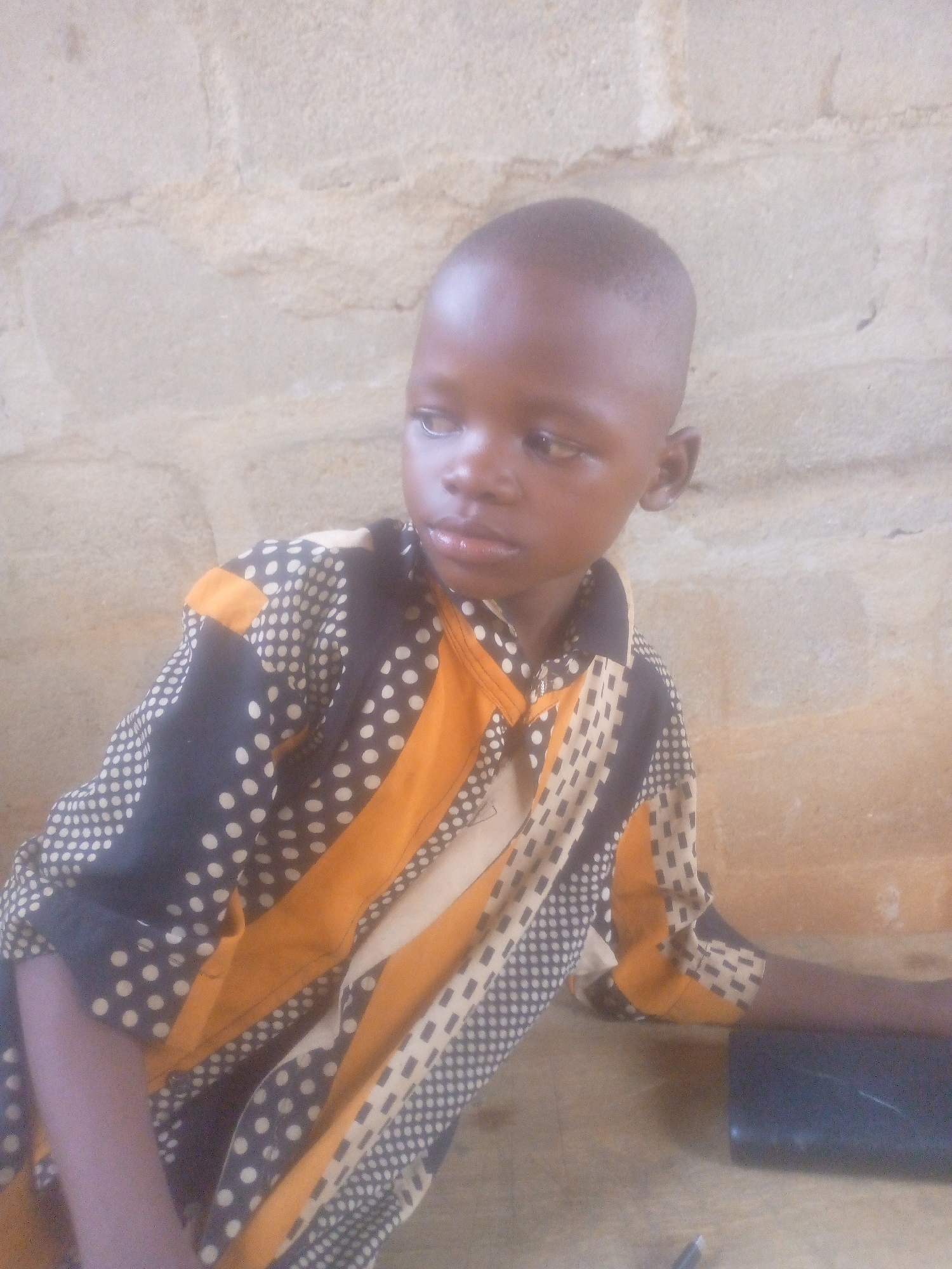 A young boy is shown in a close-up, slightly high-angle shot, sitting at a table in an indoor environment. He has short, close-cropped hair and is wearing a patterned shirt with stripes of black polka dots, yellow, and white. He is looking off to the side. On the table, to the right, is a dark object and a pen. The background is a textured, light-colored wall. The location is Unknown, Unknown.