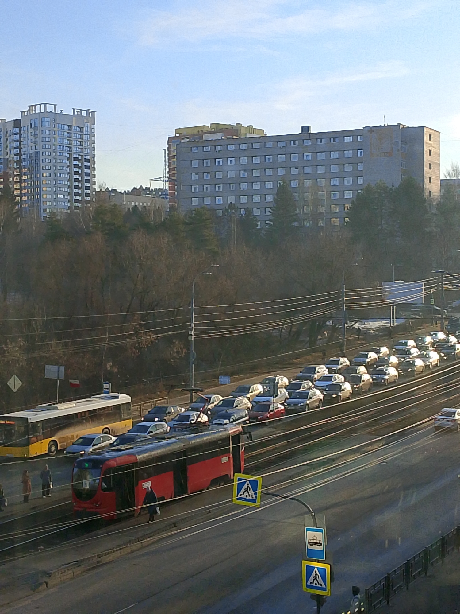 The image shows an urban scene in Izhevsk, Russia, with a mix of residential and commercial structures.
In the foreground is a street with a red tram on the tracks and a yellow bus on the road. Both public transportation vehicles are stationary. People are standing near the tram.
A line of cars is visible on the road, indicating traffic congestion. Above the street and cars are numerous overhead power lines. Beyond the road, there is a dense area of trees and bushes.
In the background are two large buildings; one is a modern apartment block with numerous windows, and the other is a Soviet-era concrete building with a more utilitarian design. The sky is blue with some clouds.