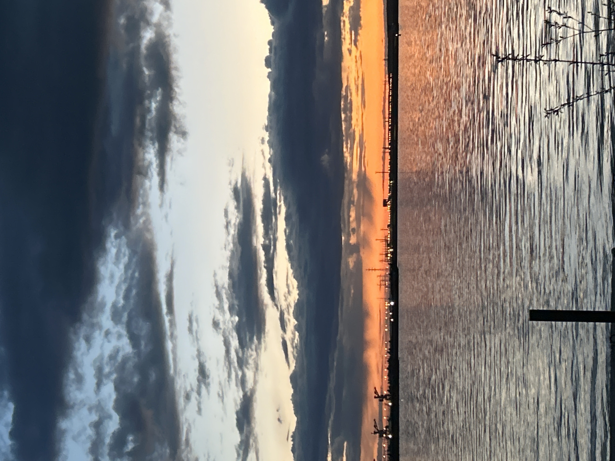 The image captures a sunset over the Kama River in Chaikovsky, Perm Krai, Russia. The sky on the left is filled with dark, layered clouds illuminated by the setting sun. The right side of the image shows the Kama River with a calm, reflective surface mirroring the sunset colors. The shoreline is sparsely populated with silhouettes of industrial structures and utility poles. In the foreground, on the right, is a darkened tree branch and the top of a metal structure. The overall atmosphere is peaceful with a contrast between the natural beauty of the sunset and the industrial presence on the horizon.