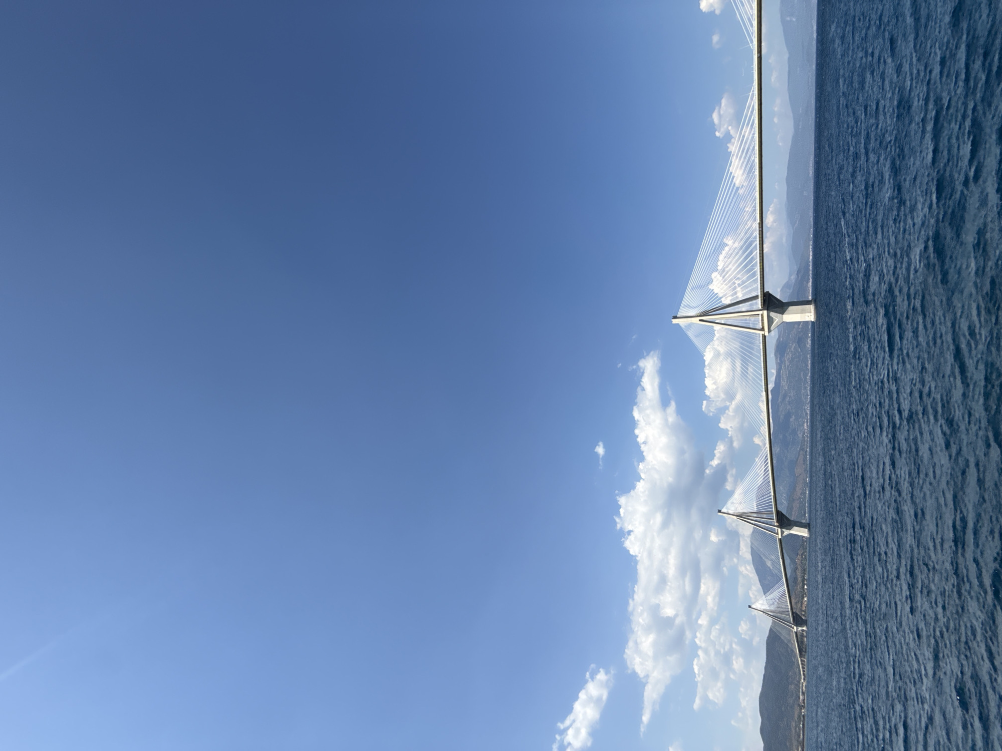 The image depicts a cable-stayed bridge spanning a body of water. The bridge is the primary object, with its tall pylons and numerous cables stretching across the frame. The water is a deep blue, with a textured surface indicative of waves or ripples. In the background, mountains rise up, some covered in greenery and others appearing rocky. The sky is a clear, bright blue with a few scattered white clouds, suggesting fair weather. The sun appears to be high in the sky, casting light and creating some highlights on the water and the bridge. The perspective is from the water level, looking towards the bridge and the distant landscape. No people are visible in the image. The scene conveys a sense of scale and engineering, highlighting the grandeur of the bridge and its natural surroundings. The Municipal Unit of Rio in Greece is known for the Rio-Antirrio Bridge, which fits this description.