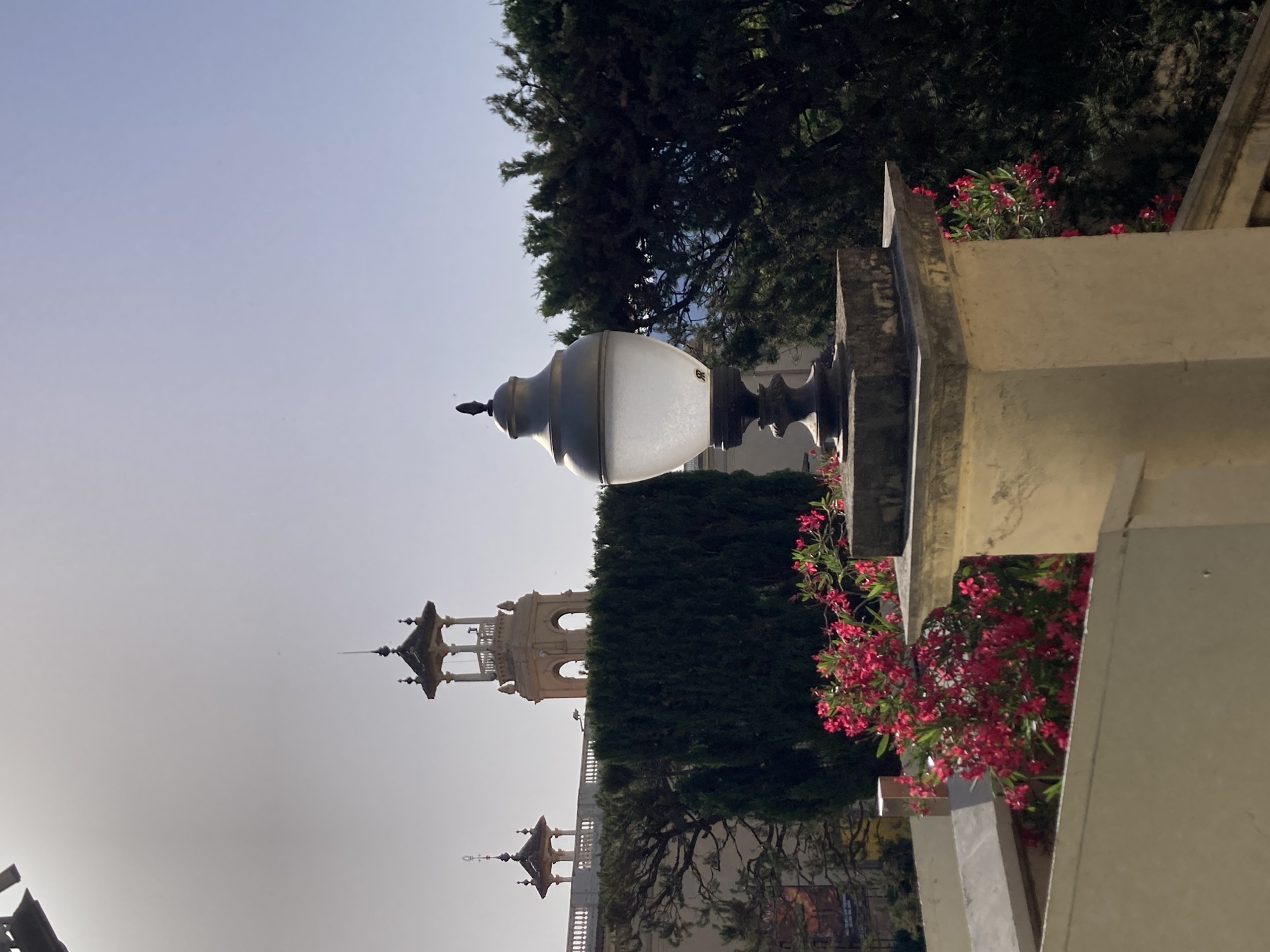The image shows a sunny day in Barcelona, Spain, with a view of a classic lamppost in the foreground. The lamppost, made of metal and glass, stands on a stone pedestal. In the background, two architectural structures with distinctive towers and decorative elements are visible. These structures appear to be part of a larger building or complex, possibly with a historical or cultural significance, and are framed by lush green trees and vibrant pink flowers. The sky is clear and blue, suggesting good weather conditions for outdoor activities. The overall scene evokes a sense of tranquility and beauty, highlighting the architectural charm and natural splendor of Barcelona.