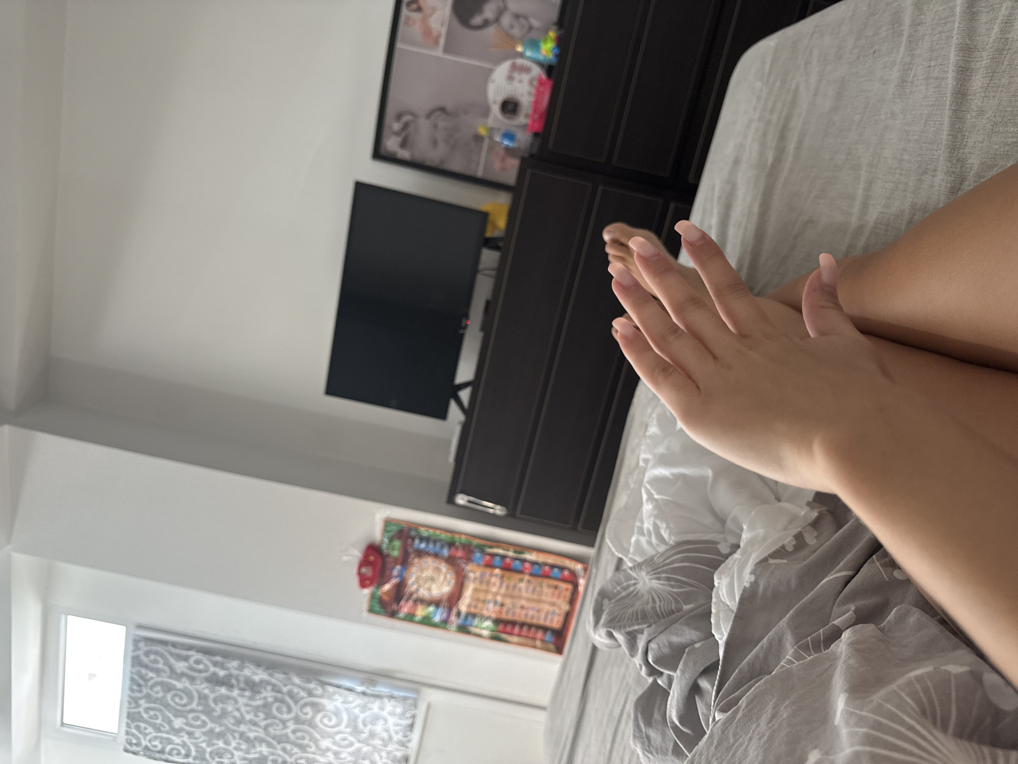 The image depicts a bedroom scene, with a person's legs and hand resting on a bed. The bed is covered with grey and white bedding. In the background, there are two dark dressers, a television, and a framed collage of photographs. A colorful textile art piece is hanging on the wall above the left dresser, and a patterned curtain is visible next to a window on the left side of the frame. The overall impression is that of a domestic, lived-in space. The image does not contain any discernible landmarks or features that would allow for specific identification of its location within Chișinău, Moldova.