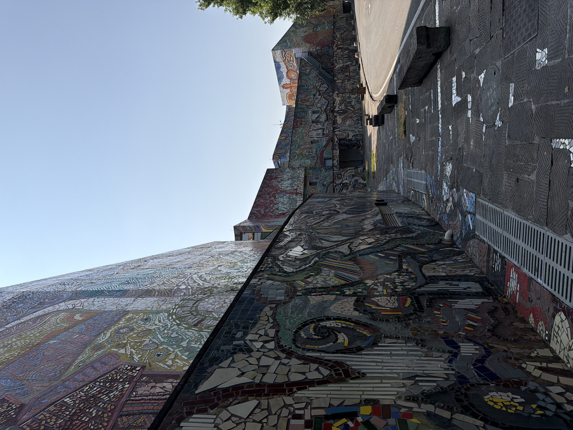 The image captures a sunny outdoor scene with a clear blue sky. The focal point is a large, multi-story building extensively decorated with colorful mosaic tile murals. The building exhibits an architectural style with multiple sections and varying heights, featuring large windows. A paved pathway runs along the base of the building, also adorned with mosaic tile patterns. Several dark, stone benches are placed along the pathway. To the right, a paved road curves away, bordered by a line of trees. The overall impression is one of a vibrant, art-filled public space. The location is identified as being in the Chongqing High-tech Zone, China. There are no people visible in the image.
