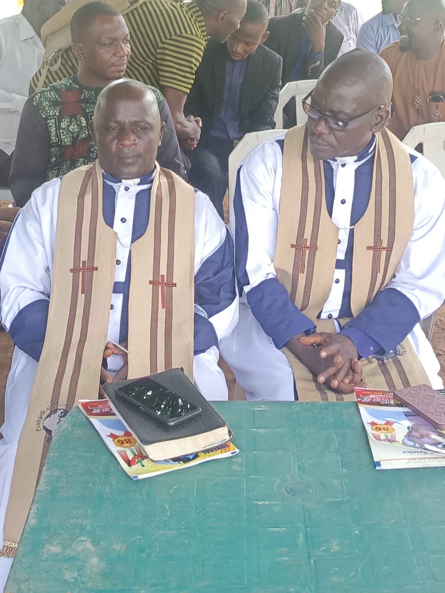 Two clergy members, dressed in white robes with blue accents and beige stoles adorned with red crosses, are seated at a green plastic table in the foreground of an outdoor or semi-outdoor gathering in Andaha, Nigeria. The man on the left, with a serious expression and balding head, looks directly ahead. A black book, likely a Bible, with a smartphone placed on top, rests on the table in front of him, along with a booklet. The man on the right, also balding and wearing glasses, has his hands clasped and is looking towards the left, indicating engagement. Several booklets, one clearly marked "SERVICE" and "80," are on the table in front of him, next to a brown patterned item.
In the background, other individuals are seated on white plastic chairs, observing the event. Some are partially visible, wearing various casual to semi-formal attire, including a striped shirt and a patterned shirt. The setting appears to be a daytime religious service or meeting, suggested by the attire of the main subjects, the Bibles, and the service booklets. The light is bright, indicating a sunny day. Partial text "Evangelical Church" is visible on the stole of the left clergyman. The overall scene suggests a community or church event.