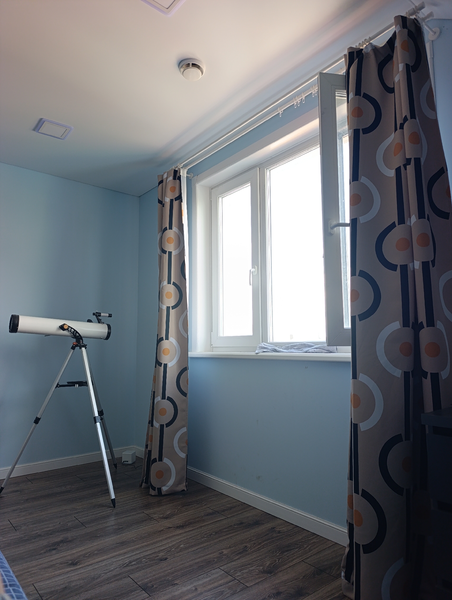 An interior shot captures a room with light blue walls and a dark wood-plank floor. A white telescope on a silver tripod stands on the left side of the room. A window with white trim dominates the right side of the frame, with one pane open. Two curtains with a geometric pattern of circles and semi-circles in beige, navy, and orange hang on either side of the window. A white curtain rod runs across the top of the window. On the ceiling, a white smoke detector is visible. The scene appears to be a domestic setting.