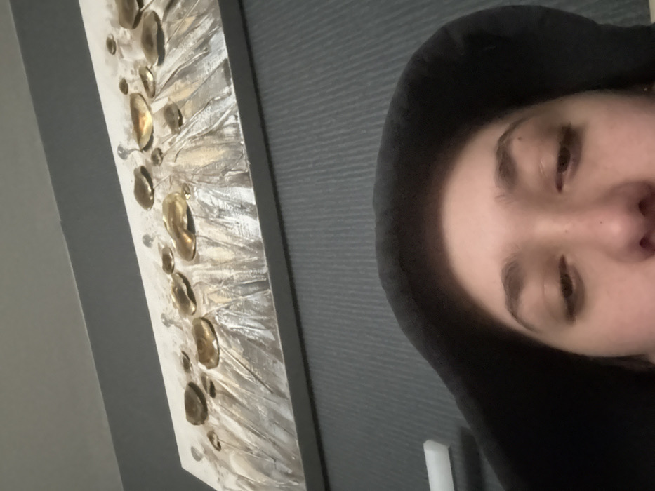 The image depicts an indoor scene focused on the upper face of an individual and a piece of wall art. A person, identified by visible eyes, eyebrows, and the bridge of their nose, occupies the lower portion of the frame. The individual wears a dark-colored hood that obscures the rest of their head and neck. Their eyes appear heavy-lidded or partially closed, directed downwards.
In the background, mounted on a wall, is a rectangular, horizontal piece of abstract art. The artwork features a light-colored, textured base with vertical, brushstroke-like elements in shades of silver and grey. Affixed to the art are approximately twelve irregular, raised, metallic gold-colored shapes, varying in size and scattered across the surface. The wall immediately below the artwork and behind the person is dark grey with a subtle, ribbed vertical texture. A white, rectangular object, possibly a light switch or fixture, is partially visible on the left against the dark grey wall. The specific location within Sofia, Bulgaria, cannot be determined from the provided visual information.