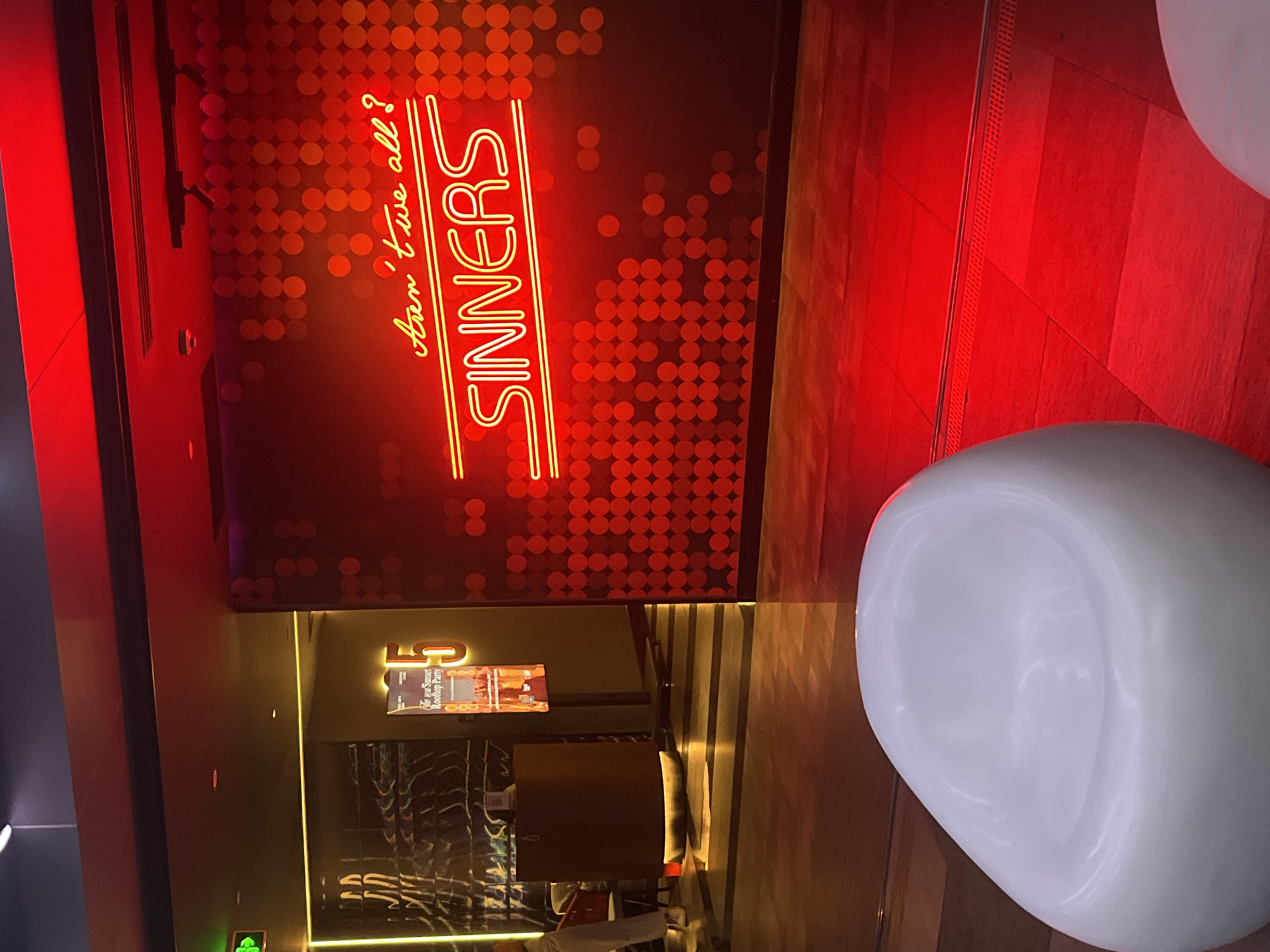 The image captures a dimly lit, modern interior, likely a bar or lounge, with a dominant red neon theme. A large, stylized sign with bright yellow neon tubing reads "SINNERS" vertically, with smaller cursive text above it that says "Aren't we all?". The wall behind the sign is decorated with a pattern of red circular lights.
In the foreground, a large, smooth, white object, possibly a chair or sculpture, occupies the lower right portion of the frame. The floor appears to be made of wooden planks, illuminated by red light. To the left, a digital display shows text that is partially obscured but includes "4F" and "Rooftop Party", suggesting a venue with multiple levels. The overall ambiance is chic and atmospheric, with a hint of playful indulgence suggested by the "SINNERS" sign. The scene is indoors, likely in the evening given the artificial lighting. The location context provided places this scene in Da Nang, Vietnam.