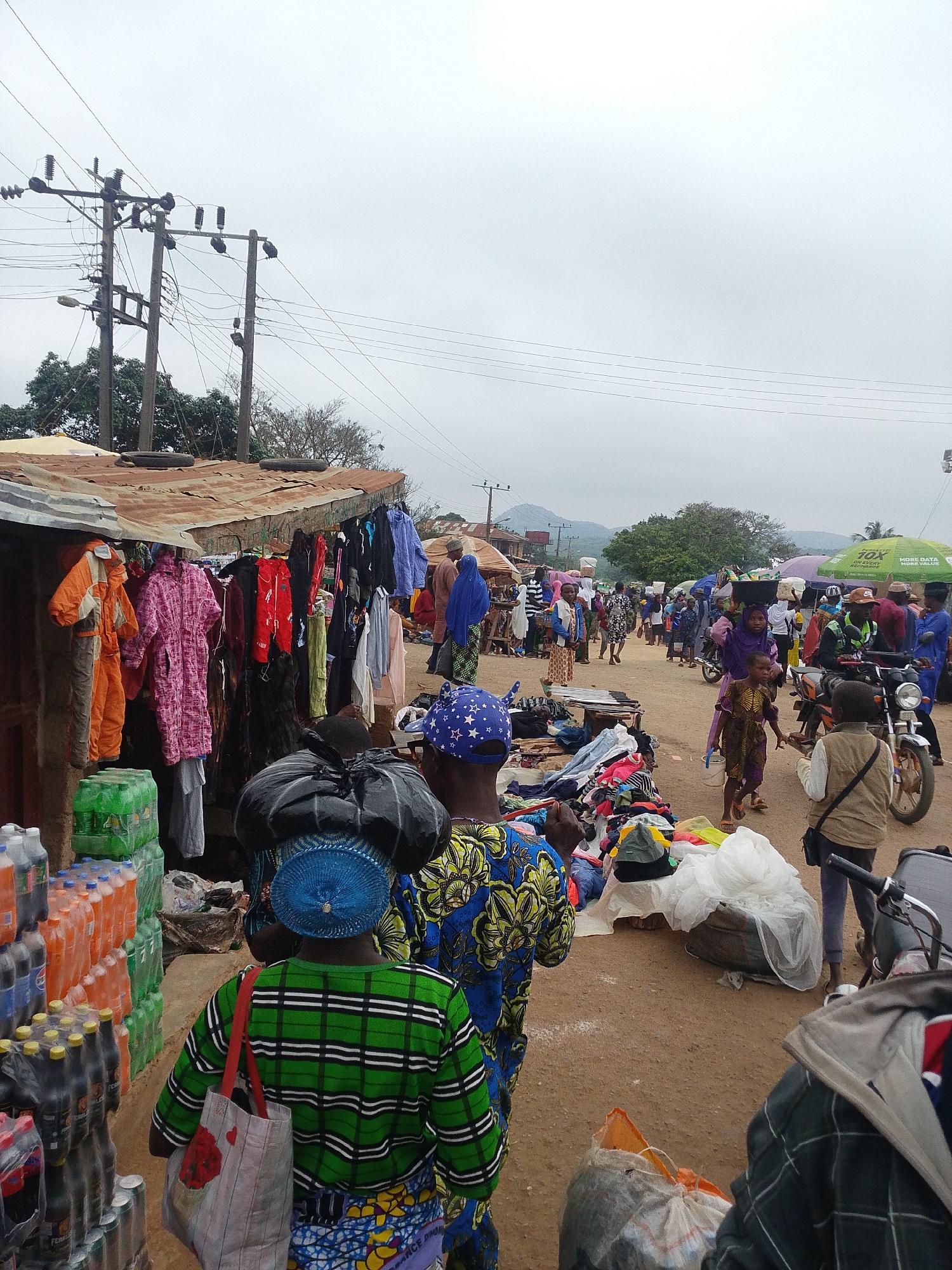 This image captures a bustling outdoor market scene in Komu, Nigeria. The foreground is dominated by a stall with various clothing items for sale, including brightly colored jackets and dresses. Stacks of bottled drinks are visible on the left, suggesting a diverse range of goods available. Several people are browsing or walking through the market.
In the midground, the market extends further, with more stalls and people. A few motorcycles are parked, indicating a common mode of transport. People are carrying items on their heads, a typical practice in many African markets. The ground is unpaved, typical of a rural or semi-urban setting.
In the background, more market activity can be seen, along with trees and distant hills under an overcast sky. Electrical poles with wires are also a prominent feature of the landscape, highlighting the presence of infrastructure. The overall atmosphere is one of vibrant commercial activity in a community setting.