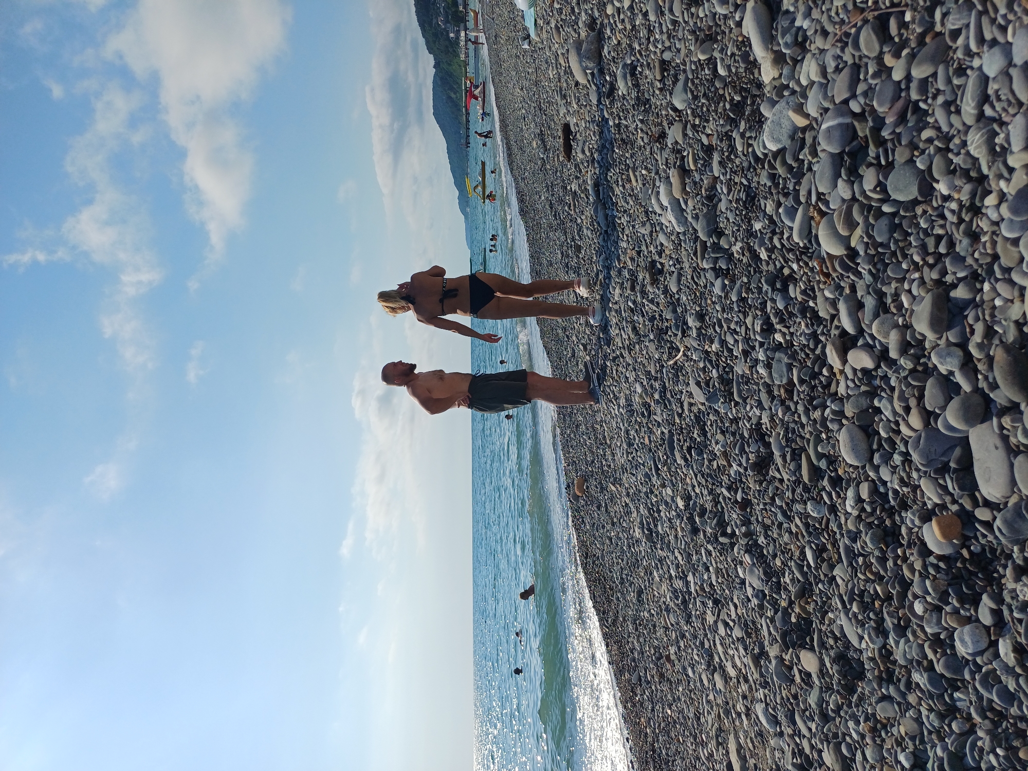 A man and a woman stand on a pebble beach with the ocean to their left and the sky above. The man, in dark shorts, stands facing the woman. The woman, wearing a black bikini, stands facing away from the man, looking out at the ocean. Her hair is blonde, and she is wearing white shoes. The pebbles on the beach are various shades of gray and brown. The ocean is a bright blue with white-capped waves. In the background, there are several other people in the water and on the beach. Mountains can be seen in the distance. The sky is a clear blue with some scattered white clouds. The scene appears to be set in a sunny location, likely during the daytime. The overall mood seems relaxed and pleasant, as one might expect on a beach vacation. The location is indicated to be Sochi, Russia.