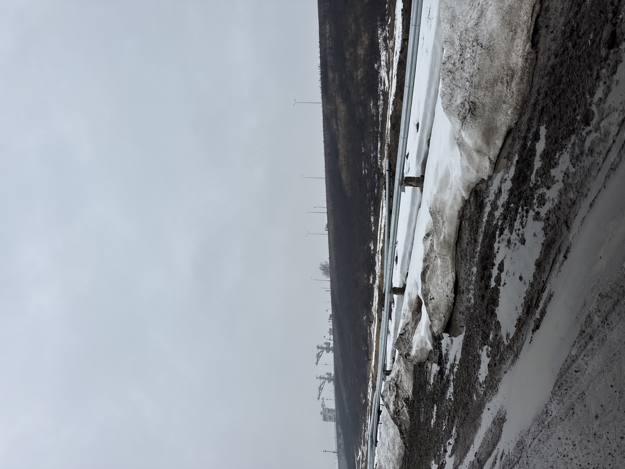 The image shows a winter scene in what is likely Chaikovsky, Russia. There's a road or pathway with melting snow and mud alongside a hillside. A metal guardrail runs along the side of the road. On the hillside, there is a line of bare trees and some industrial structures are visible in the distance. The sky is overcast.