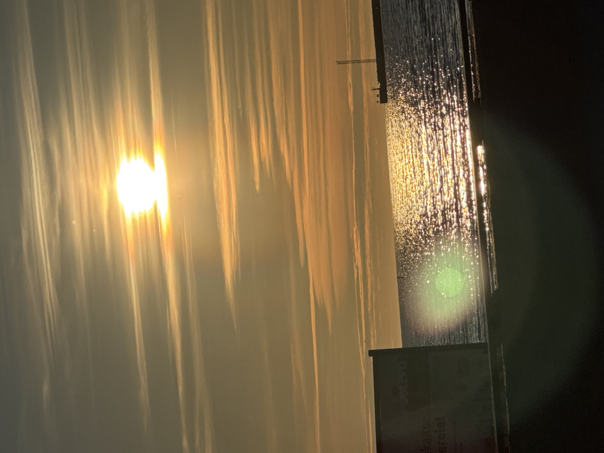 A vertical, eye-level shot captures a scenic view of a body of water, likely the Baltic Sea, at sunset in Tallinn, Estonia. The sun, a bright, white orb with a halo of golden light, is positioned in the upper center of the frame, casting a shimmering path of light across the water. Wispy, horizontal clouds stretch across the sky, tinged with orange and yellow from the setting sun.
In the distance, a long pier or breakwater juts out into the water, with a tall, slender structure, possibly a lighthouse or communication tower, visible at its end. The water itself is a deep blue, reflecting the vibrant colors of the sky and the bright sparkle of the sun.
In the foreground, on the left, a dark, rectangular object, possibly a sign or a panel, is partially visible. Some text, appearing to be in a foreign language, is faintly discernible on its surface. The lower part of the image is dominated by a dark, shadowy foreground, suggesting a shoreline or a raised platform. A faint green lens flare is present in the middle left of the image, over the water. The overall atmosphere is serene and picturesque, characteristic of a sunset over the sea.