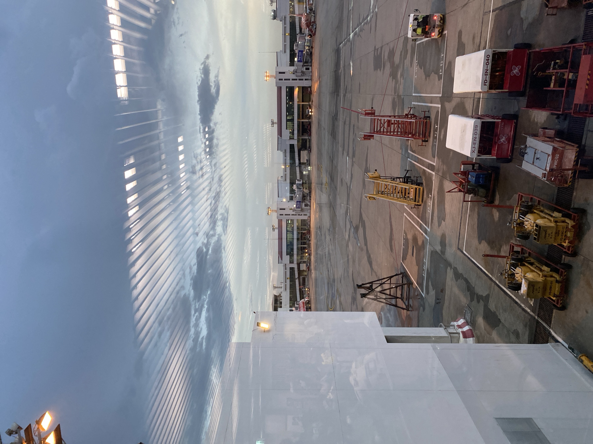 The image shows an outdoor area, likely an airport tarmac, with various pieces of ground support equipment. There are several vehicles, including baggage carriers and other motorized equipment. The vehicles are painted in a variety of colors, including red, white, and yellow.
In the background, a building with multiple levels is visible. The image also shows an aircraft painted with a prominent "asia.com" logo, suggesting the location is in Asia.
The presence of airport support equipment and the aircraft suggests that this scene is at Suvarnabhumi Airport (BKK) in Bangkok, Thailand. The reflection off the windows is from the window blinds, and the weather is cloudy.