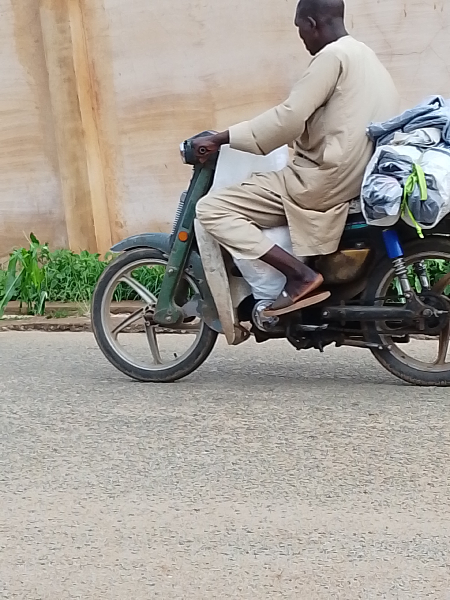 A man is riding a green motorbike on an asphalt road, with a large bundle of white and gray items tied behind him. The man is wearing a light beige robe, pants, and brown sandals. The background features a beige wall with some plants growing along the base. The location is likely in Jos, Nigeria, based on the appearance of the man and the style of the motorbike, as well as the vegetation and architecture.