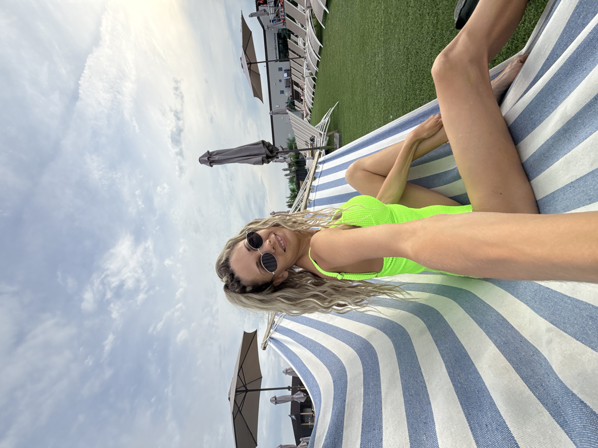 A woman with blonde, braided hair and sunglasses is lying in a hammock with blue and white stripes. She is wearing a neon green swimsuit. Her right arm is extended, holding the camera, which suggests she is taking a selfie. The hammock is suspended outdoors on a grassy area with rows of lounge chairs and umbrellas in the background. The sky is overcast with some lighter clouds. The location is identified as Rusanovo, Russia.
