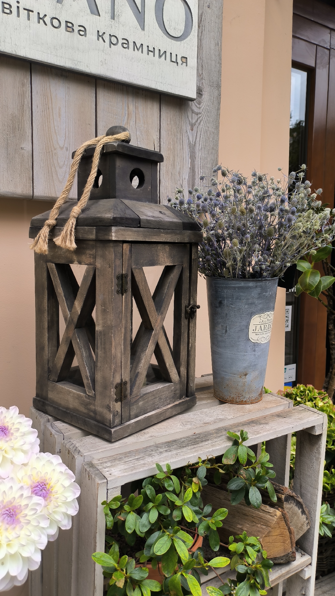 A rustic wooden lantern sits on a weathered wooden crate in front of a wall with Ukrainian text. To the right of the lantern, a distressed metal pot holds a bouquet of dried blue thistle flowers. In the foreground, a cluster of light purple and white dahlias is visible. The scene is likely an outdoor display for a shop or cafe in Lviv, Ukraine, given the presence of Ukrainian text.