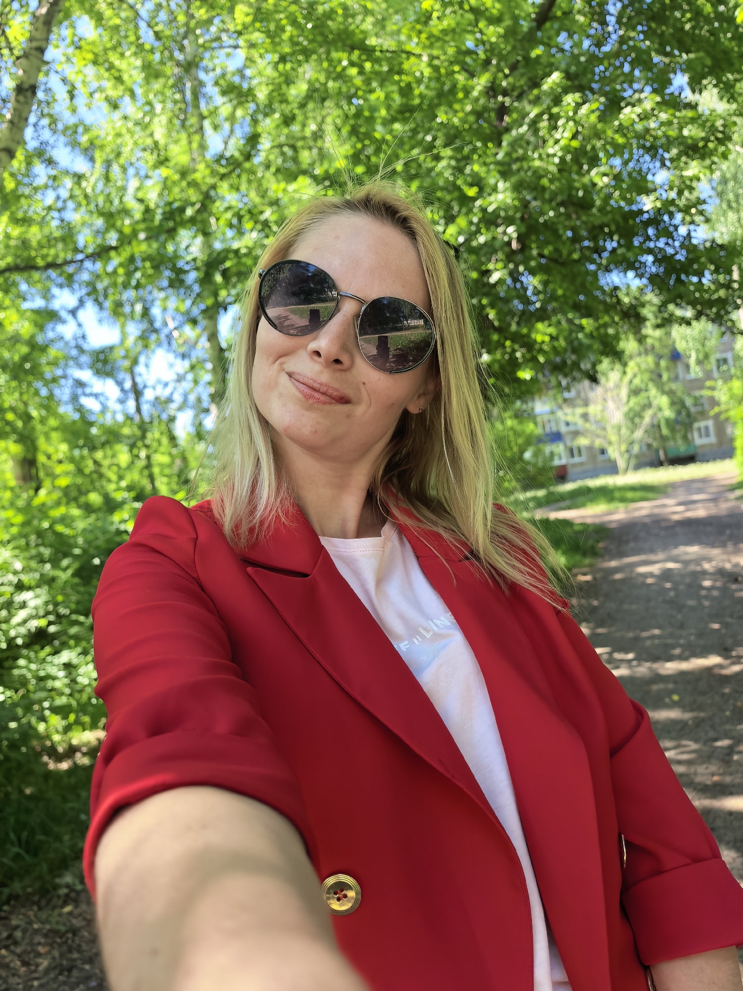 A blonde woman wearing a red blazer and sunglasses is taking a selfie. She is outside in what appears to be a park or green space with a path, trees, and a building in the background. The environment suggests she is in the city of Sterlitamak, Russia. The sunlight is bright, indicating it is likely daytime.