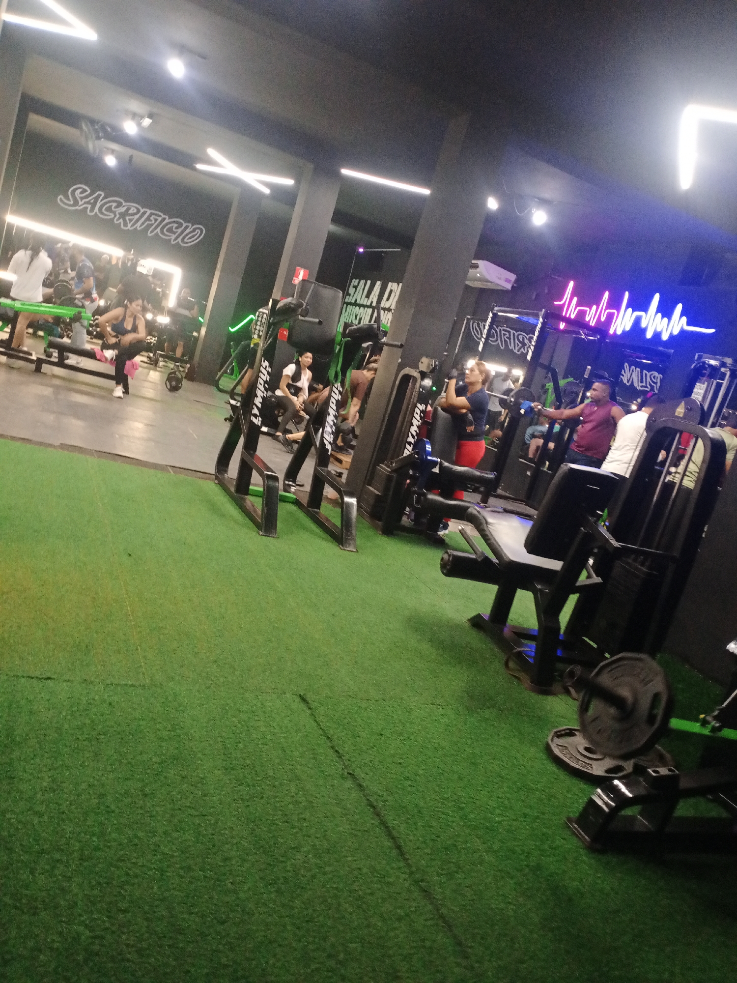 The image displays the interior of a modern gym in Valle de La Pascua, Venezuela, characterized by dark walls and ceiling, and a bright green artificial turf floor.
Numerous individuals are present, engaged in a typical workout session. On the left, several people are seen lifting weights and using benches, reflected in a large mirrored wall. A woman in a blue top and black leggings is stretching or resting near a weight bench in the mid-ground. Towards the right, other gym-goers are utilizing various exercise machines; a person is seated on a leg press-type machine in the foreground, and another is performing an exercise with a cable machine.
Notable details include the prominent artificial lighting. Linear white neon lights are mounted on the ceiling and walls, some forming an 'X' shape. A striking pink and purple neon sign, resembling an electrocardiogram (ECG) heartbeat pattern, adorns a dark wall section on the right. Opposite this, reflected in the mirrored wall, is a white neon sign that reads "SACRIFICIO." Other visible text includes "SALA D..." (likely part of a larger sign like "SALA DE MUSCULACIÓN") on a wall, and branding such as "OLYMPUS" and "shadow.W" on the black gym equipment. The overall atmosphere is energetic, typical of an active fitness center, with the artificial lighting suggesting it could be day or night.