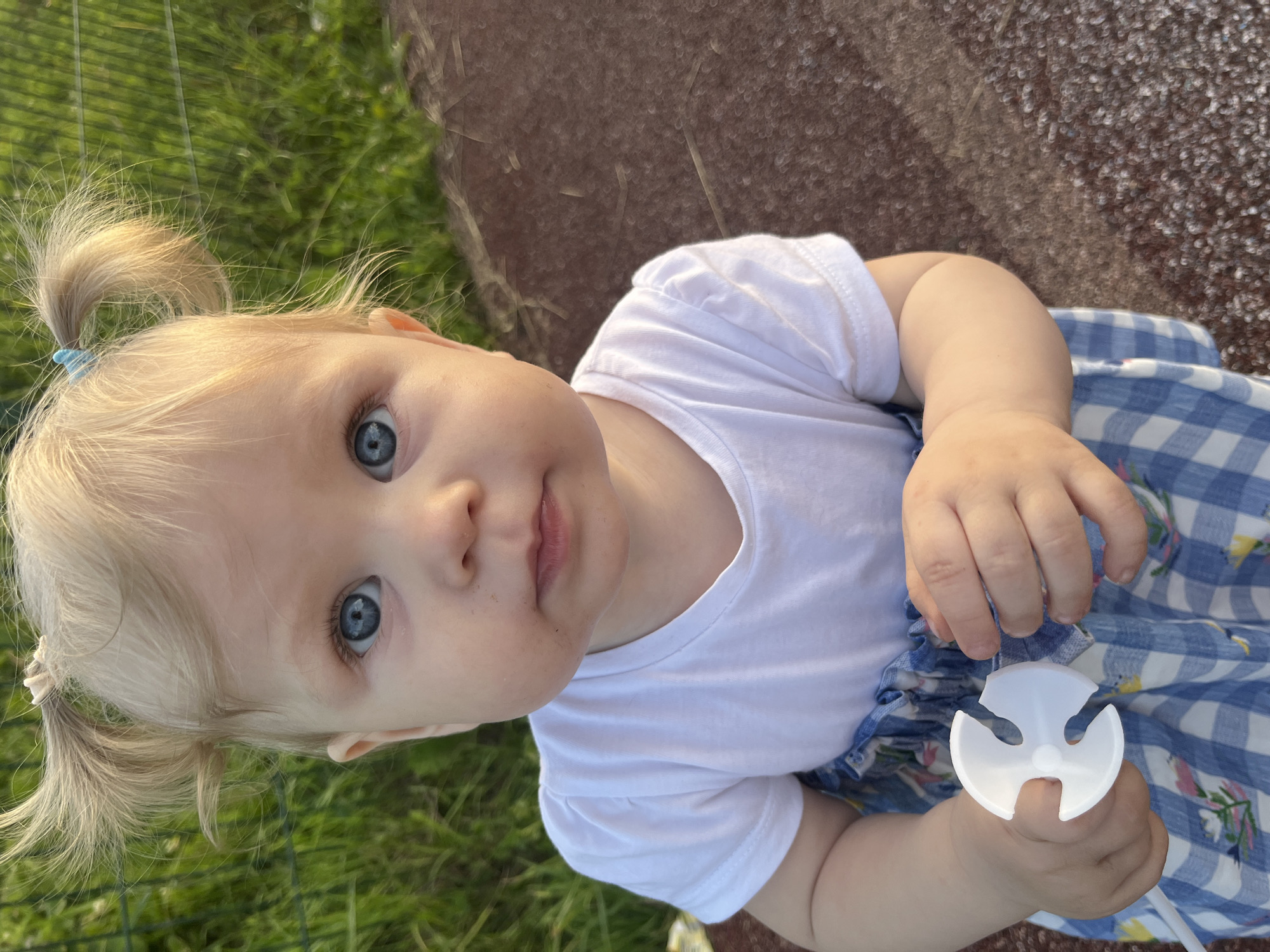 A blonde baby with blue eyes is pictured outdoors. The baby is wearing a white t-shirt and a blue and white checkered dress. The baby is holding a white plastic object in their hands. The background consists of green grass and a brown textured surface. The location is the city of Фоки, Russia.