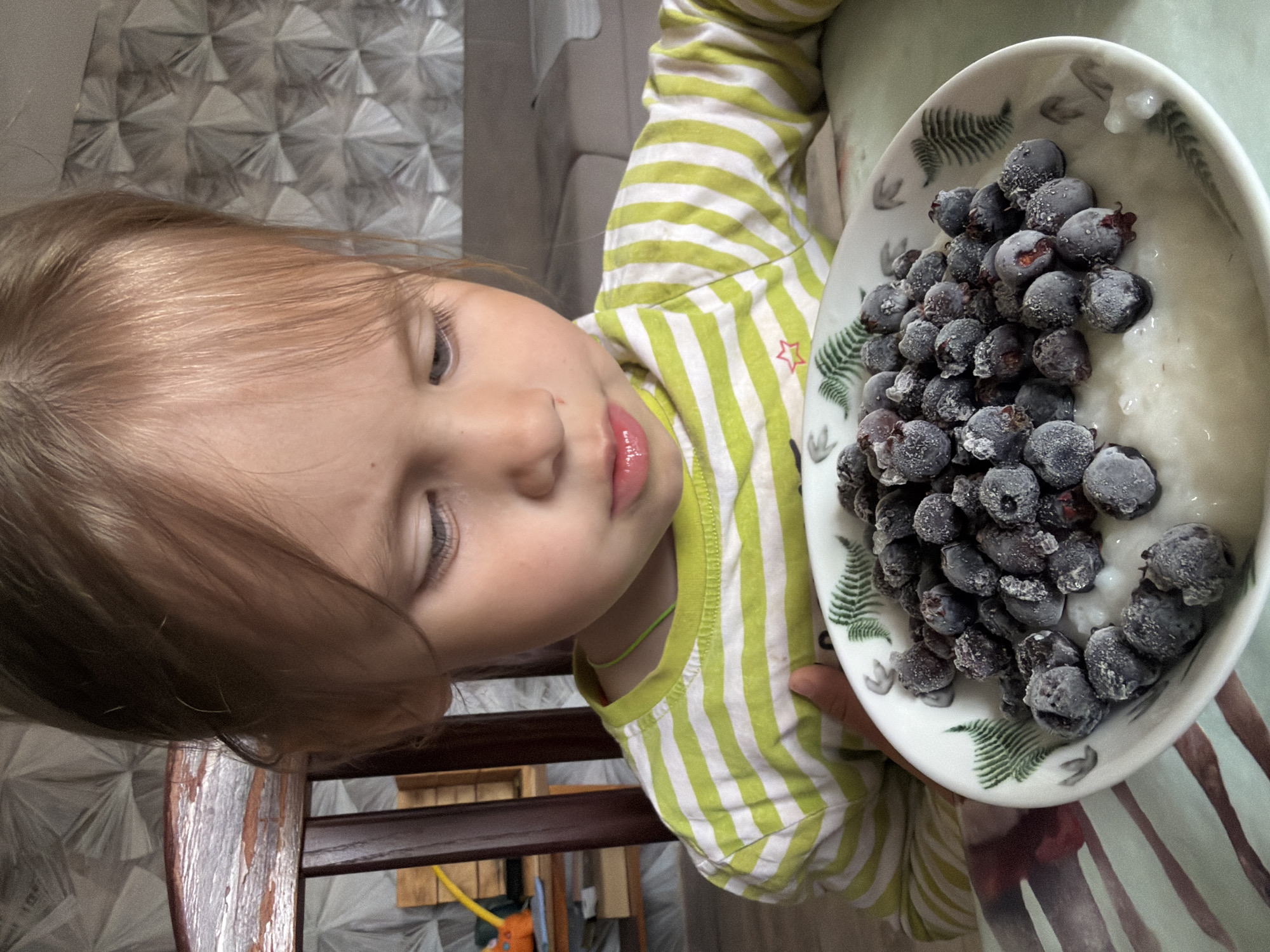 An indoor, brightly lit photograph captures a young child focused on a bowl of food, likely in a home setting in Chaikovsky, Russia. The child, with light brown or blonde hair, is seen from the chest up, positioned on the left side of the frame with their head tilted. They are wearing a long-sleeved shirt with alternating light green and white horizontal stripes, featuring a small red star emblem near the shoulder. A thin, light green string or necklace is visible around their neck. The child's eyes are closed or cast downwards, and their lips are slightly parted, conveying a serene or contemplative expression, possibly anticipating the meal.
The child is holding or reaching for a white bowl, which occupies the right side of the image. The bowl is decorated with green fern or leaf patterns along its rim. It contains a large quantity of dark blue berries, which appear to be frozen due to a visible frosty coating, resting on a creamy white substance, possibly yogurt, porridge, or a similar milky base.
The background features a wall covered in a distinct, light-colored wallpaper with a repeating geometric, possibly three-dimensional, pattern. Part of a dark wooden chair back is visible in the lower foreground. The overall scene suggests a peaceful, daytime moment during a meal or snack time. No explicit text is visible in the image beyond the small red star on the child's shirt.