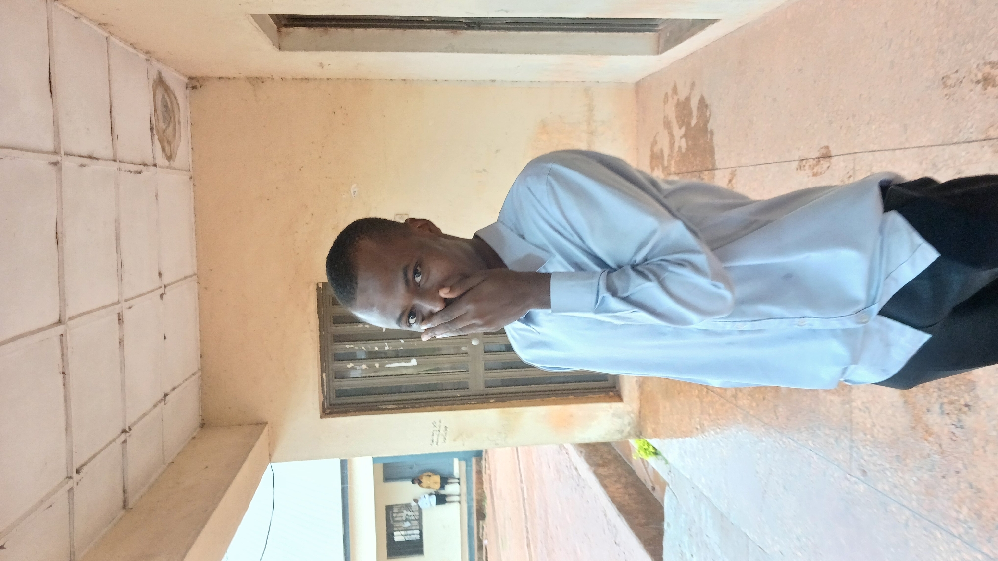 A man stands indoors near a window with metal bars, partially covering his mouth with his hand. He wears a light blue, long-sleeved shirt and black pants. In the background, visible through a lower window, are two people standing near a building. The interior features beige walls and tile work. The image was captured in the city of Jos, Nigeria.