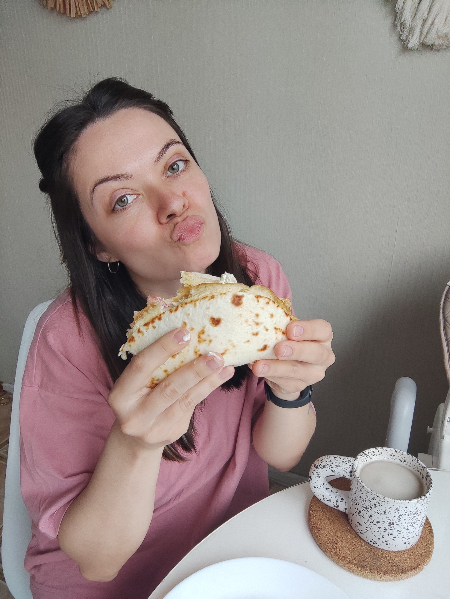 A woman is indoors, holding a partly eaten flatbread in both hands while puckering her lips. She has dark brown hair, light skin, and green eyes, and is wearing a pink shirt and silver hoop earrings. She has a black watch on her left wrist. To the right of her, on a white table, is a white speckled mug sitting on a brown cork coaster, presumably containing a beverage. A white plate is partially visible near the mug. The wall behind her is a neutral color. A macrame decoration is partially visible in the top left corner of the image, and part of a high chair is visible in the background on the right. The city and location are Unknown, Unknown.