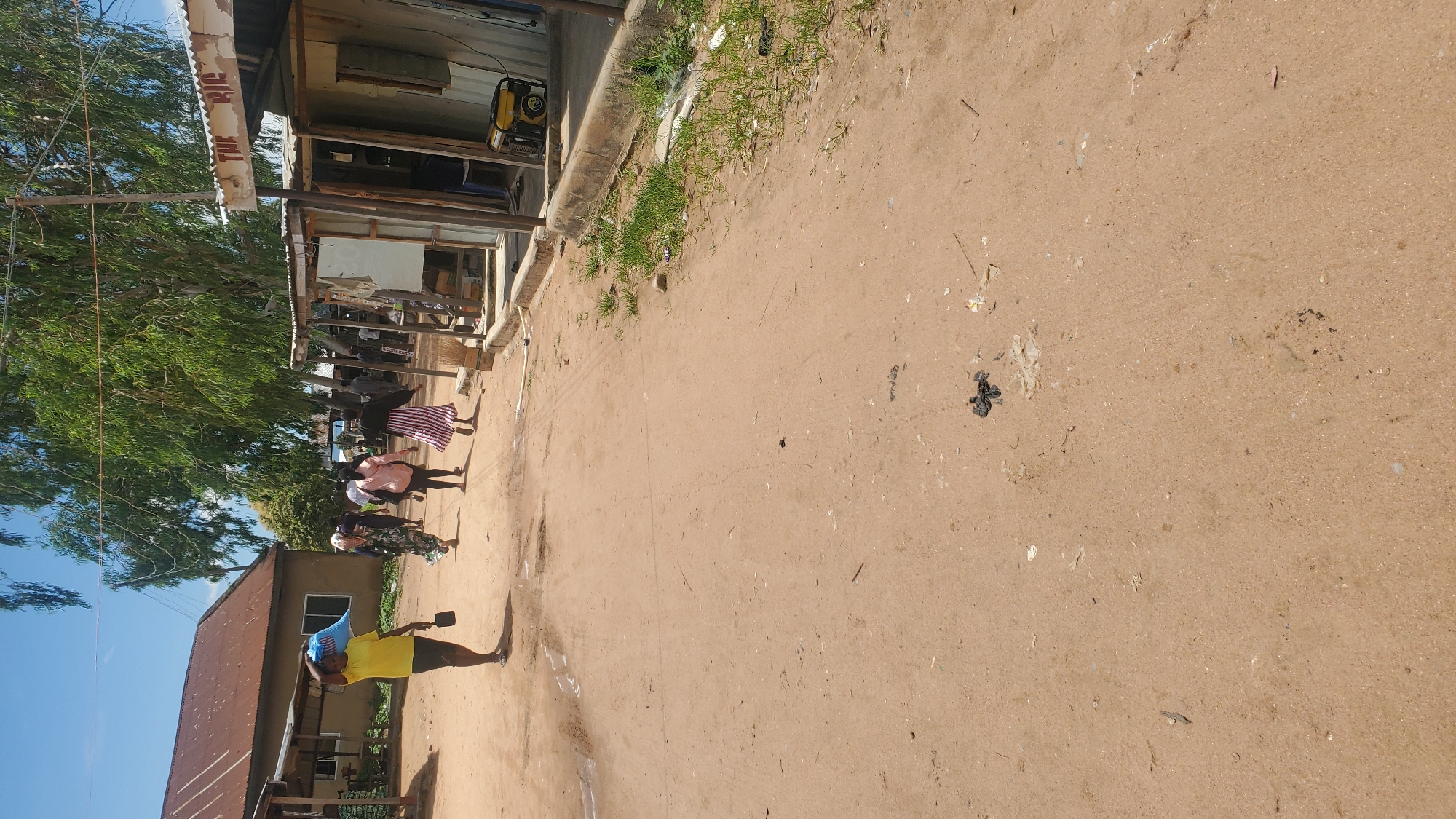 In Jalingo, Nigeria, a dirt road runs through a street lined with informal structures and trees. A woman in a yellow shirt and black shorts carries a blue bag on her shoulder and a dark object in her right hand as she walks away from the camera. Several other people, including women and children, are visible walking down the road. One woman wears a patterned dress, another a pink top and black pants, and a third a striped dress. The buildings on either side of the road appear to be constructed from corrugated metal and wood. A generator is visible outside one of the structures. The ground is dusty and scattered with debris. The sky is blue with some clouds.