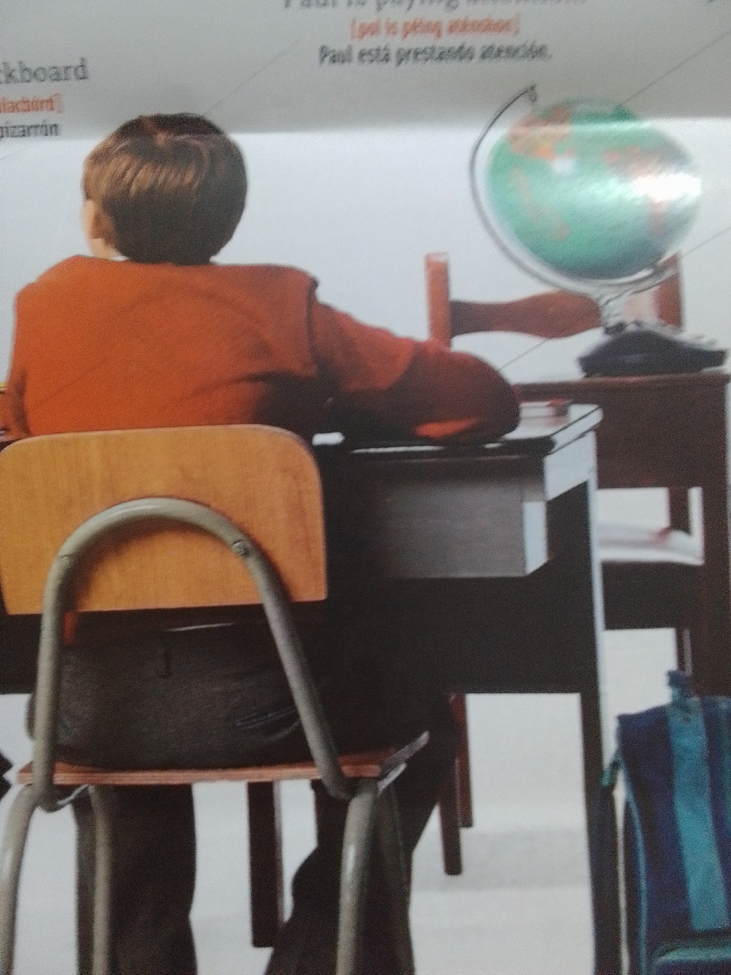 The media file shows a young boy from behind, wearing an orange shirt and black pants. He is sitting at a wooden desk with a drawer. On the desk, there is a globe. The scene appears to be a classroom or study area. The boy is facing away from the viewer and towards the desk and globe. The lighting suggests it is daytime. There is text visible in the background, partially obscured. One line of text appears to say "blackboard" and another "pizarrón". Further text reads "Paul is paying attention" and "Paul está prestando atención". This suggests the context is related to learning or education. The location is given as La Tagua, Venezuela.