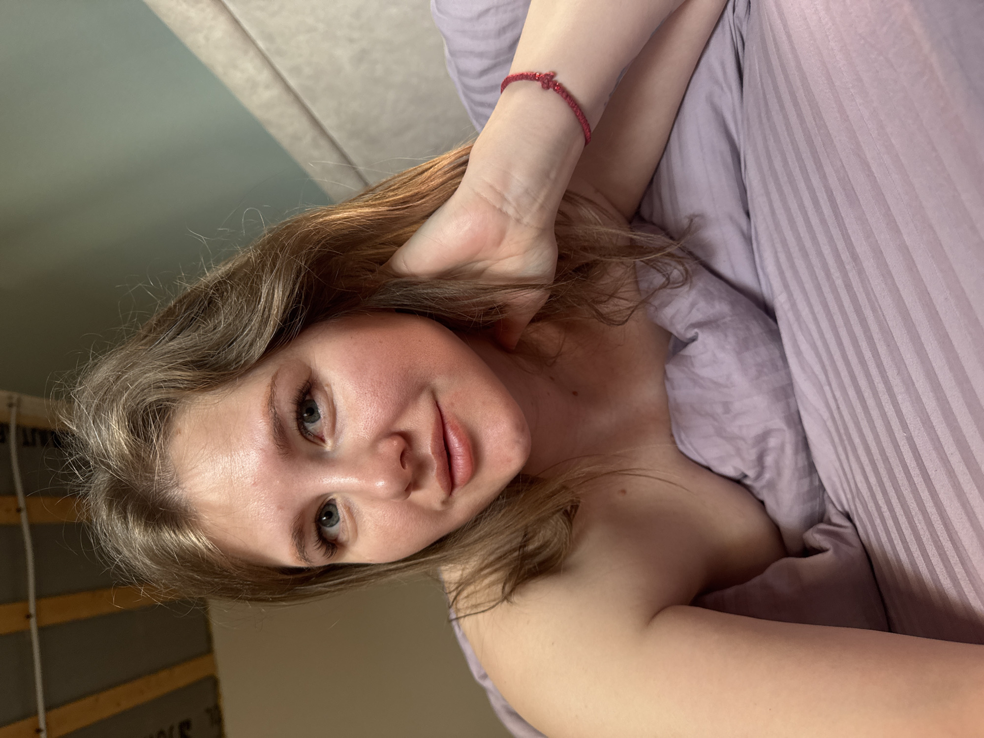 A young woman is lying in bed, looking directly at the camera. She has fair skin, light brown hair, and blue eyes. Her left hand is raised, with her fingers touching her hair near her temple. She is wearing a red string bracelet on her left wrist. She is partially covered by a light purple duvet. The headboard of the bed is visible in the background, along with a wall. Based on the prompt, the location is inferred to be Gornyi (Dvurechenskoe S/P), Russia.