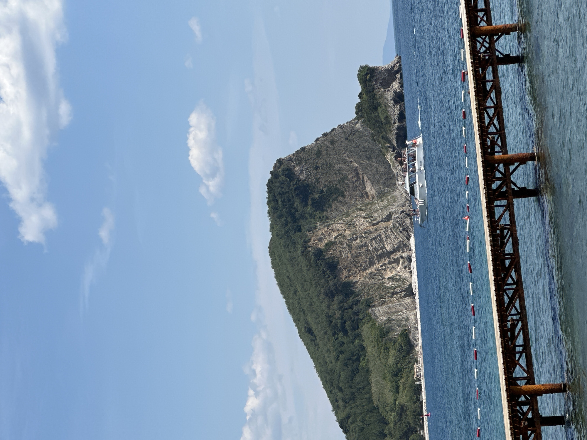 A landscape featuring a body of water, a pier, and a mountainous shoreline, likely near Budva, Montenegro. The image captures a pier with rusted metal supports extending into the water, marked by a line of red and white buoys. Adjacent to the pier, a mountain covered in dense vegetation rises from the sea, with visible geological stratification. A boat is docked alongside the shore with several people on board. Above, a blue sky is scattered with several puffy clouds.