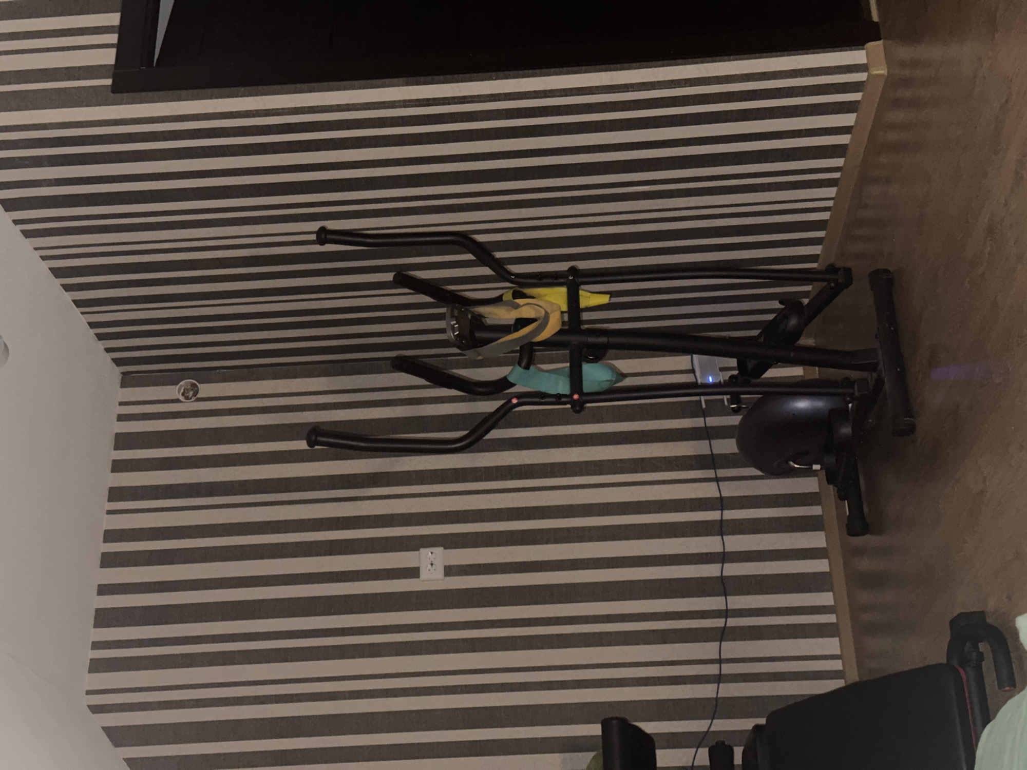 In a room with striped wallpaper and a wooden floor, an elliptical exercise machine stands prominently. Attached to the machine are a teal strap, a yellow strap, and a gray strap. To the left of the elliptical, a black weight bench is partially visible. An electrical outlet with two sockets is mounted on the wall. A dark brown door is located on the right side of the frame. The image captures a scene likely within a residential setting in Shcheglovo, Russia.