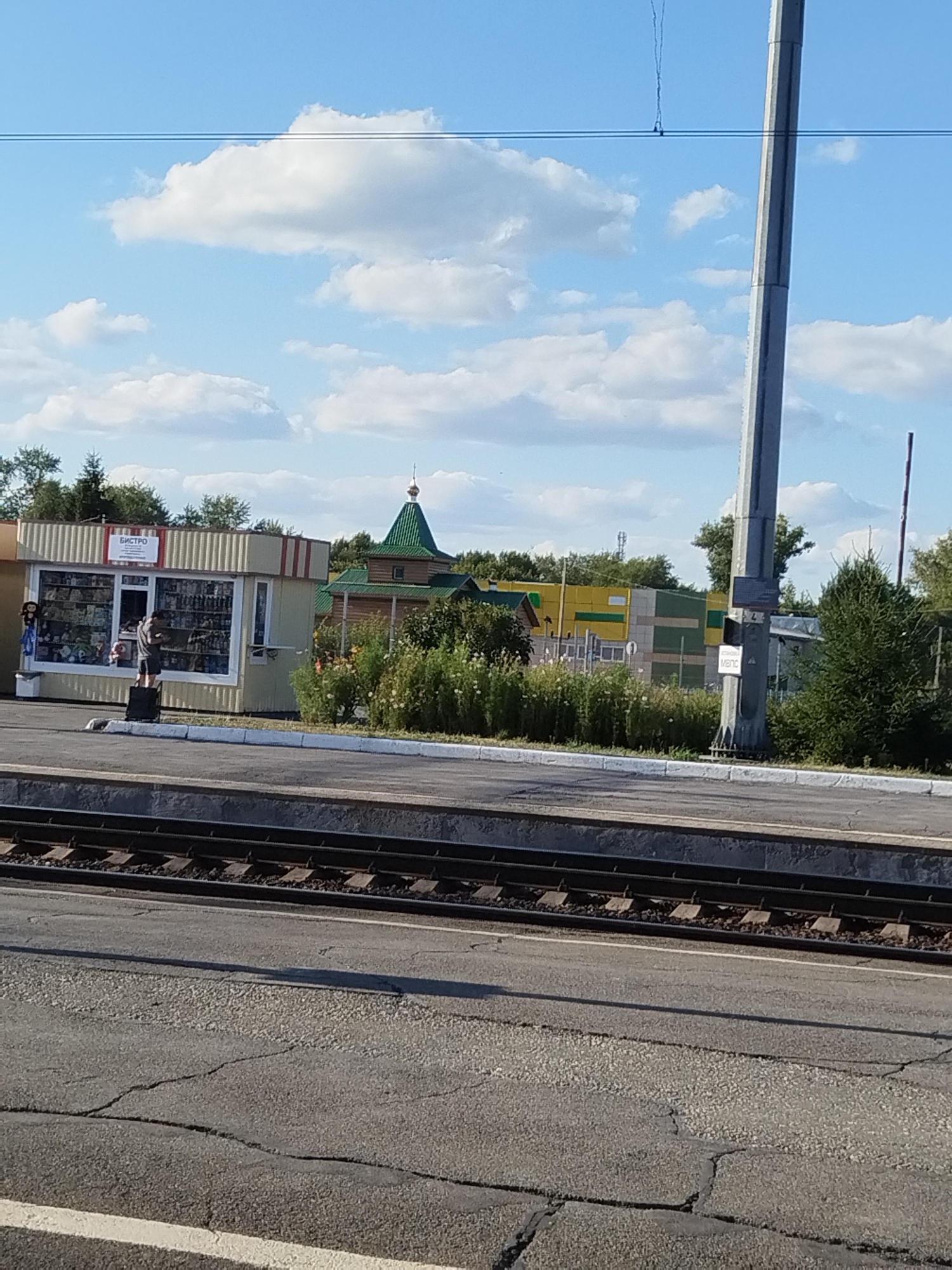 A daytime, medium shot captures a scene at a train station in Barabinsk, Russia. In the foreground, a section of train tracks is visible, with ballast and ties supporting the rails. Adjacent to the tracks is a paved area with visible cracks and some lighter-colored aggregate surfacing. Beyond this, a raised concrete platform runs along the tracks.
Further back, a small, light-colored kiosk with a sign reading "БИСТРО" (Bistro) stands. The kiosk has large windows displaying various items, and a person in a gray t-shirt is seen standing outside, appearing to be interacting with the kiosk. To the right of the kiosk, a low-lying building with a green, multi-tiered roof, reminiscent of a church or chapel, is situated. It is constructed of wood. Behind the kiosk and the chapel-like structure, there is dense green foliage and trees.
In the background, a tall, grey concrete pole, likely part of the station's infrastructure, dominates the right side of the frame. Signs are affixed to this pole. Further back, a long, low building with horizontal stripes of yellow and green is visible, along with other structures and trees. The sky is blue with scattered white clouds.