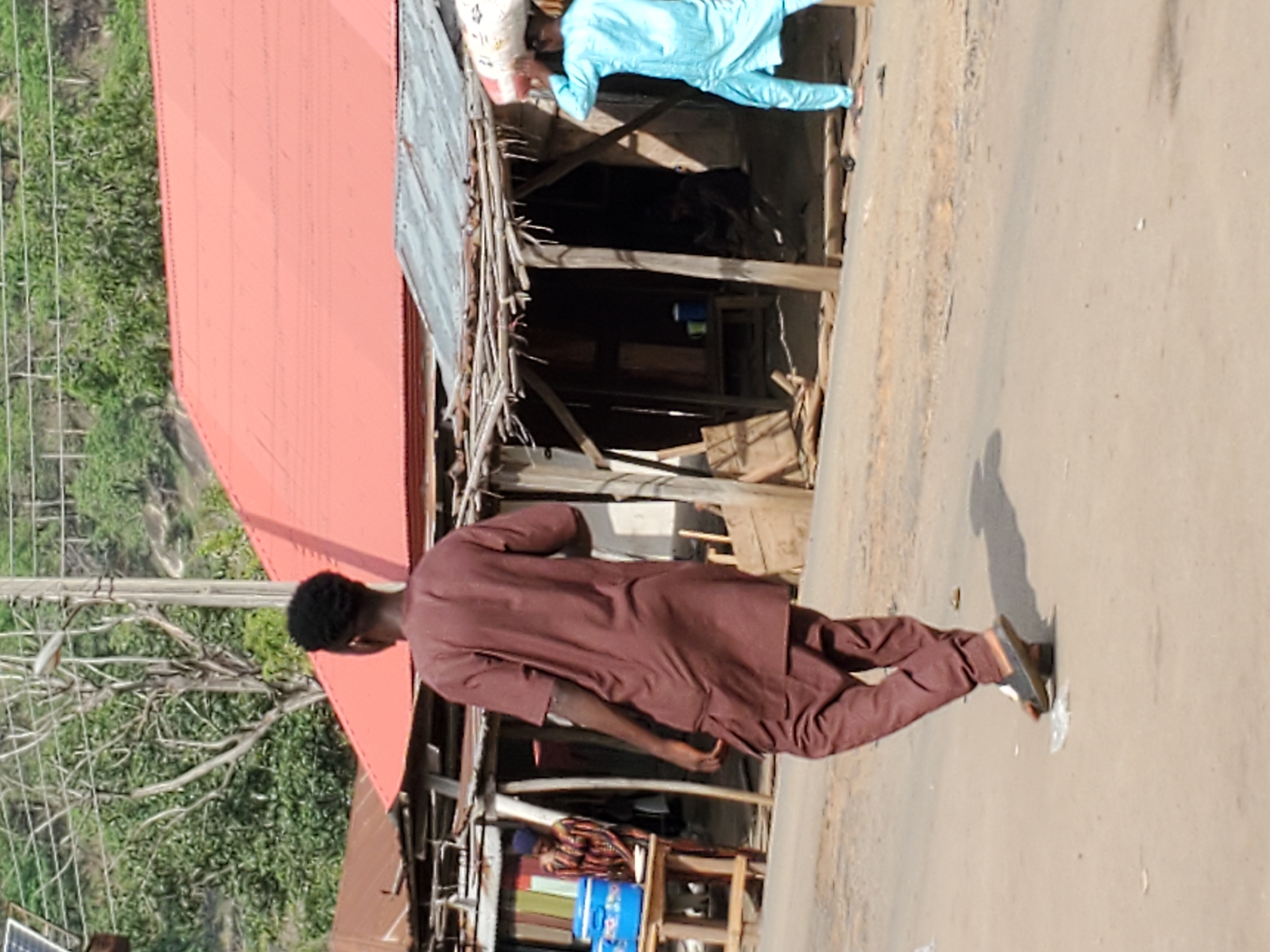 This image, originally oriented vertically but depicting a horizontal scene, captures an outdoor environment in Mararaba, Nigeria, under bright daylight.
In the foreground, a person dressed in a maroon or dark brown traditional tunic and trousers walks away from the viewer on a light-colored, possibly unpaved road. A distinct shadow extends from their right, indicating a strong light source, likely the sun, from the left.
The mid-ground features a series of simple, semi-permanent structures or stalls, constructed from wooden frames and corrugated iron sheets. One prominent structure on the left has a bright red corrugated iron roof. To the right of the walking individual, near these stalls, another person dressed in a light blue tunic and trousers is seen bending over. This individual appears to be engaged in an activity such as inspecting or arranging items inside a stall, whose interior seems dark but shows some wooden shelving and blue containers.
The background reveals lush green vegetation and what appears to be a metal fence, suggesting a natural or undeveloped area beyond the immediate roadside. A portion of a solar panel is visible in the bottom-left corner of the frame. The scene conveys typical daily activity in what appears to be a market or local commercial area. The bright lighting suggests it is daytime, likely mid-morning to late afternoon, with clear weather. No discernible text is visible in the image.