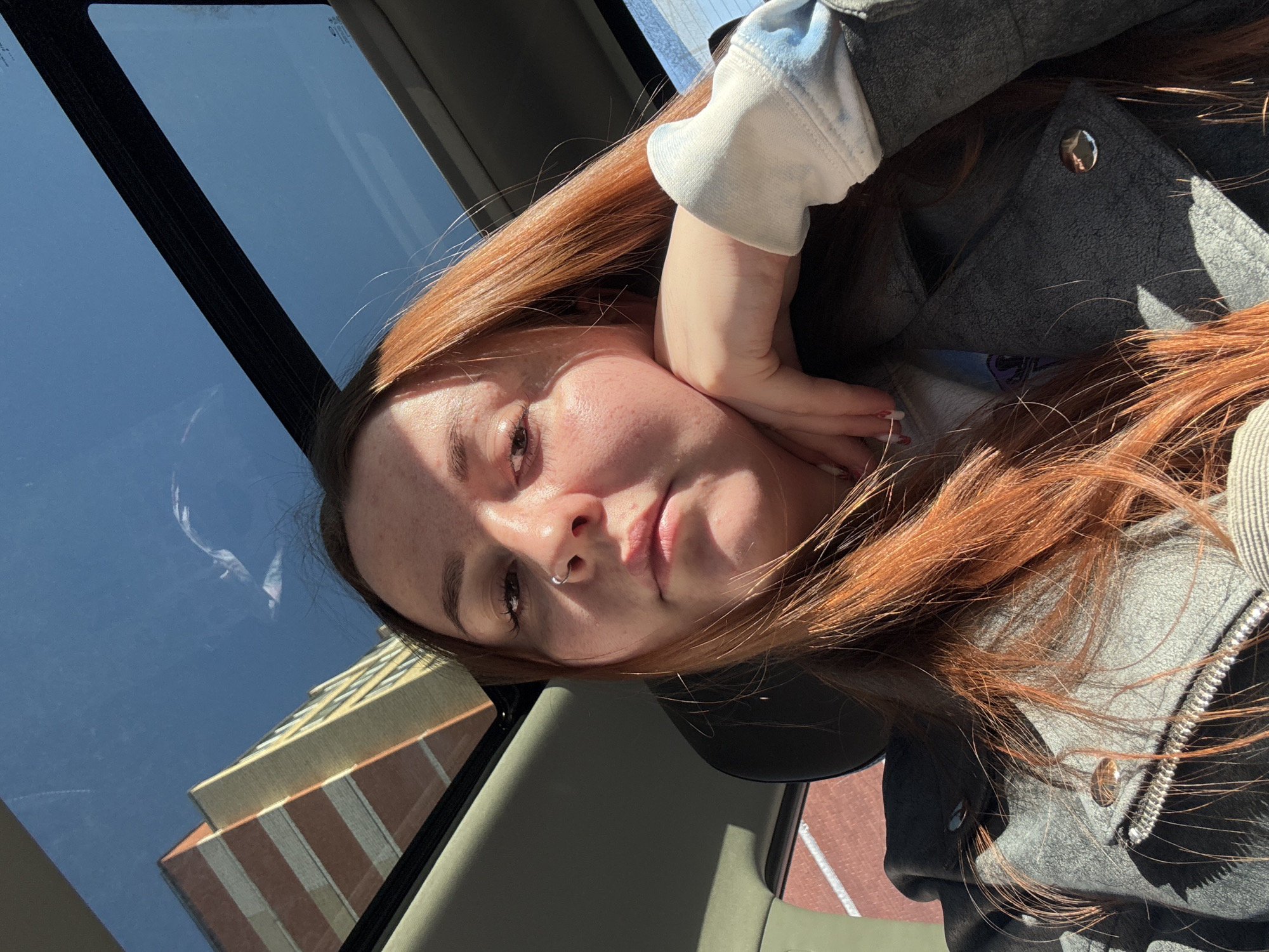 A young woman with long, reddish-brown hair is inside a vehicle, possibly a car, with a visible sunroof. She is resting her cheek on her hand. She has a nose ring. Outside the vehicle, a building with a light yellow and red brick facade is visible. The location appears to be in an urban area, but without additional visual context, it is not possible to confirm the precise location as Izhevsk, Russia.