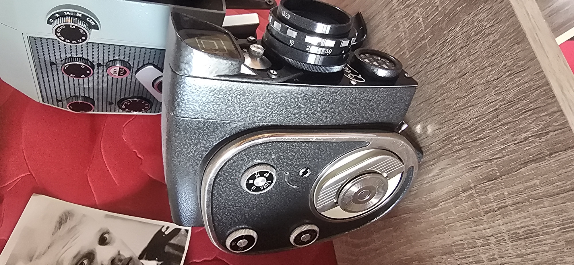 This image shows a vintage camera, possibly a Super 8 film camera, placed on a wooden surface. The camera is gray and has a textured finish. It features several dials and knobs, some with visible inscriptions. A prominent lens is attached to the front. Next to the camera, a black and white photograph of a person, potentially an actor or celebrity, is partially visible. The background includes a red, textured surface, possibly upholstery, suggesting the camera is on furniture. The setting appears to be indoors, with artificial lighting. The camera has various settings labeled, including numbers and Cyrillic text which appears to say "Е.А." and "ГОСТ". Another dial has "15", "2", "30" and "4329" written on it, as well as "V.I.R". The overall mood is one of nostalgia and appreciation for vintage technology. The camera itself is the primary subject, with the photograph adding a layer of historical context. The image is taken from a high angle, looking down at the camera.