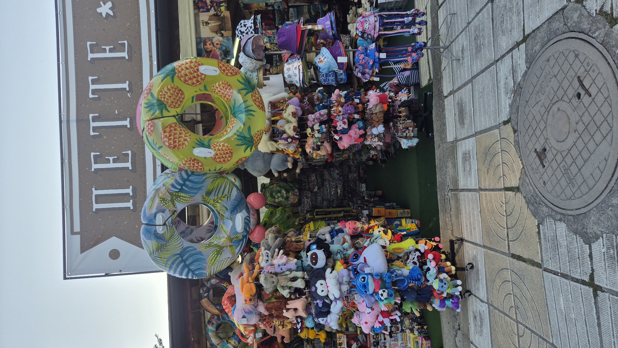 The image shows a street-level view of a market stall in Kiten, Bulgaria, featuring an assortment of merchandise, primarily stuffed animals and inflatable rings. Above the stall, a sign displays the word "ПЕТТЕ" in large, stylized letters, with a small asterisk to the right. Two inflatable rings are prominently displayed at the top of the stall; one has a blue and green tropical leaf pattern, and the other is yellow with a pineapple design.
The stall itself is laden with numerous stuffed toys of various characters and sizes, hanging from racks and displays. Below the inflatable rings, to the right, a rack of hats and sun visors is visible, in a range of colors and patterns. The stall is set up along a paved sidewalk, with a manhole cover in the foreground. The overall impression is that of a vibrant, colorful market offering novelty items, likely catering to tourists. The background suggests a building facade with some posters or prints. The lighting indicates it is daytime, possibly late afternoon. There are no people clearly visible in the image.