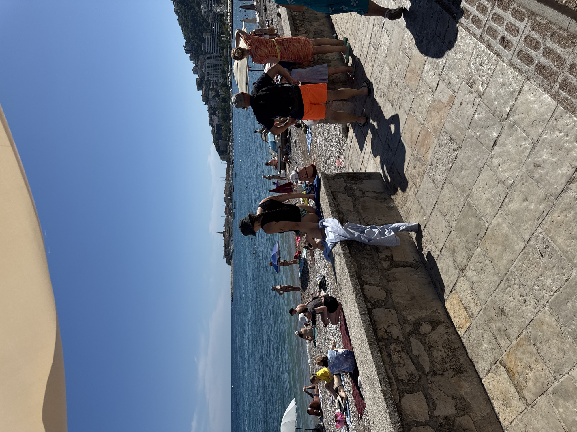 This image depicts a sunny day at a pebble beach in Budva, Montenegro, with the Adriatic Sea and a coastal cityscape in the background. In the foreground, a stone wall runs horizontally, with a tiled walkway in front of it. A light blue shirt is draped over the wall.
Several people are visible on the beach. Near the foreground, a man wearing a dark t-shirt and orange shorts stands facing away from the camera, with a backpack. Beside him, a woman in a patterned orange dress and a young girl in a light blue dress are also standing. Further down the beach, a woman wearing a black swimsuit and hat is seated on the wall, facing the sea.
The beach itself is covered in grey pebbles and populated with sunbathers. Some are lying on towels, while others are seated. Beach umbrellas are spread across the sand, providing shade. In the water, a few boats are visible, and further out, the hilly coastline of Budva is lined with buildings and hotels. The sky is clear and blue.