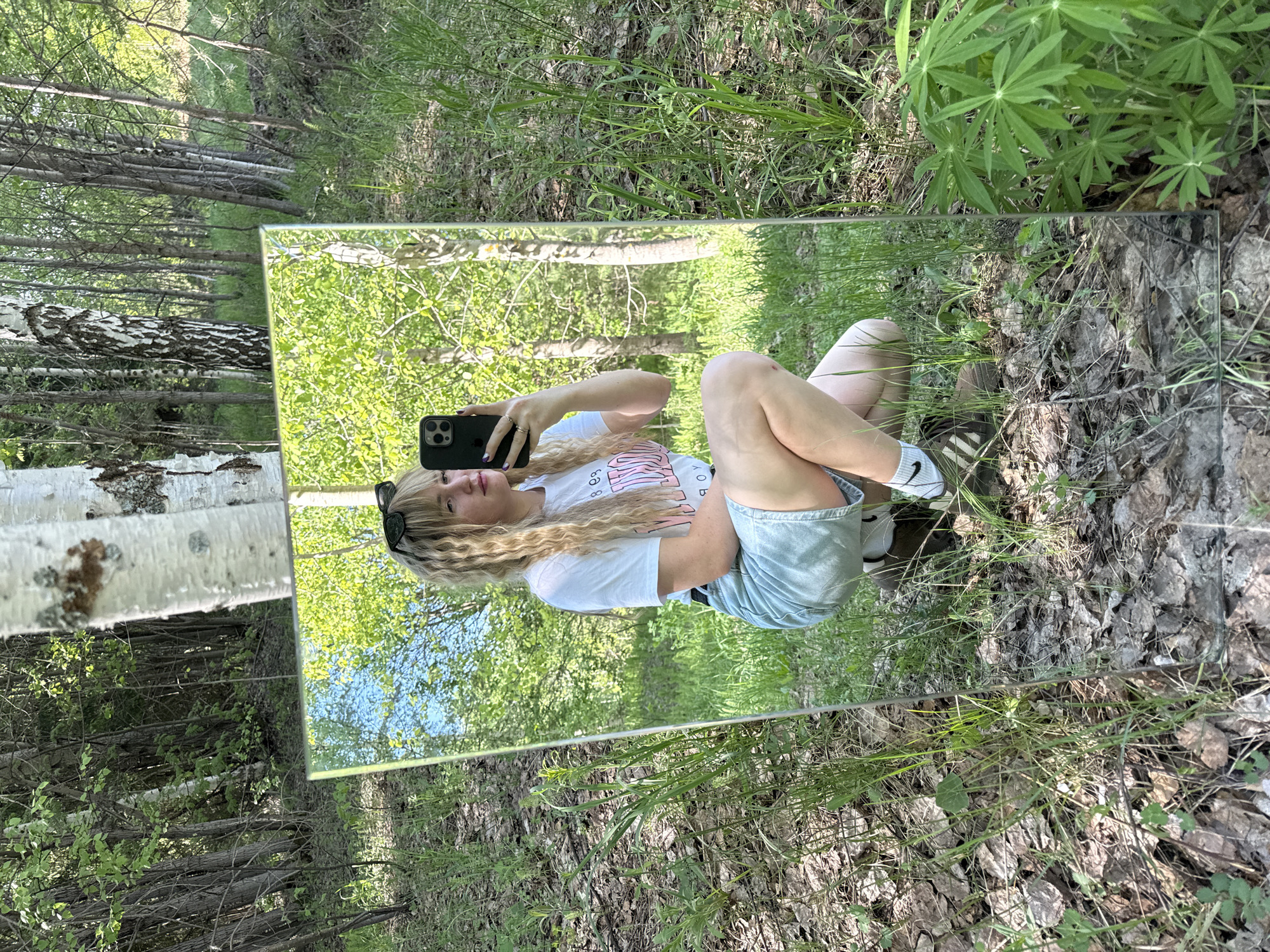 A woman in a white T-shirt and denim shorts is taking a selfie in a mirror placed in a forest near Марковский, Russia. The mirror reflects the surrounding trees and ground vegetation. The ground is covered in dry leaves and grass. The trees in the background include birch trees.