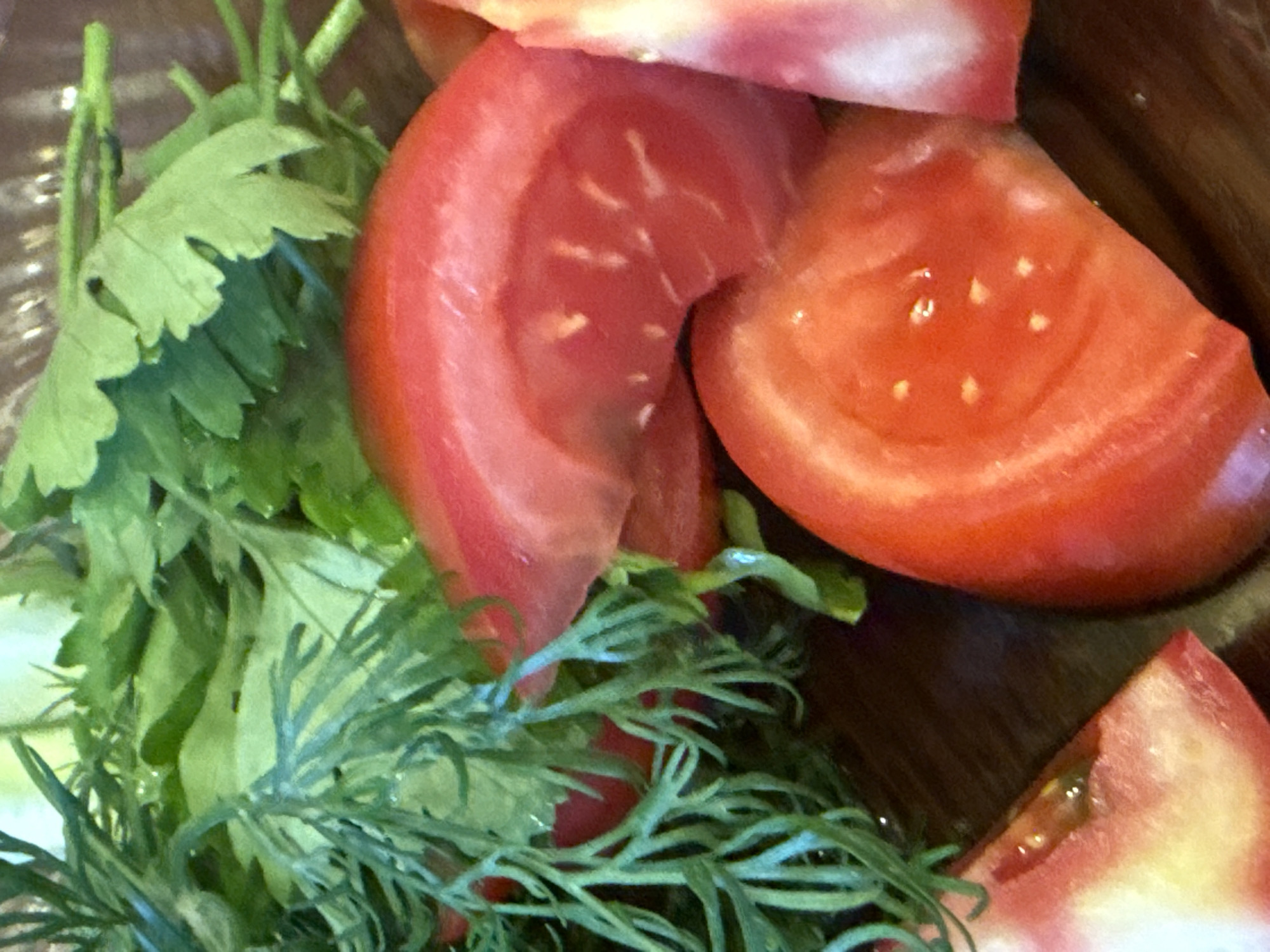 This media file is a close-up, eye-level shot of sliced tomatoes and fresh herbs, likely a salad or garnish. The subjects are the bright red slices of tomato, revealing their inner texture with seeds and pulp, and the vibrant green leaves of parsley and dill. The tomatoes appear to be ripe and juicy, with a slightly glossy surface. The herbs are fresh, with delicate fronds of dill and larger, serrated leaves of parsley.
The scene is tightly cropped, focusing on the textures and colors of the food. There is no discernible activity or event taking place, as the food is stationary. The lighting appears to be natural or well-lit artificial light, highlighting the freshness of the ingredients. The background is a dark, out-of-focus surface, possibly a wooden bowl or plate, which provides a subtle contrast to the bright colors of the food. There are no visible people, text, or specific location cues that would indicate the time of day, weather, or precise setting. The overall impression is one of fresh, healthy food preparation.