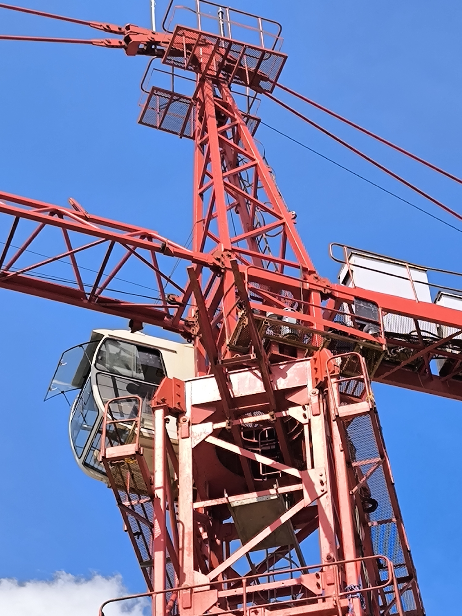 A close-up shot shows the upper section of a red construction crane against a clear blue sky. The crane's intricate lattice structure is visible, including the operator's cab on the left with an open window. Wires and cables extend from the top of the crane. The location is Unknown, Unknown.