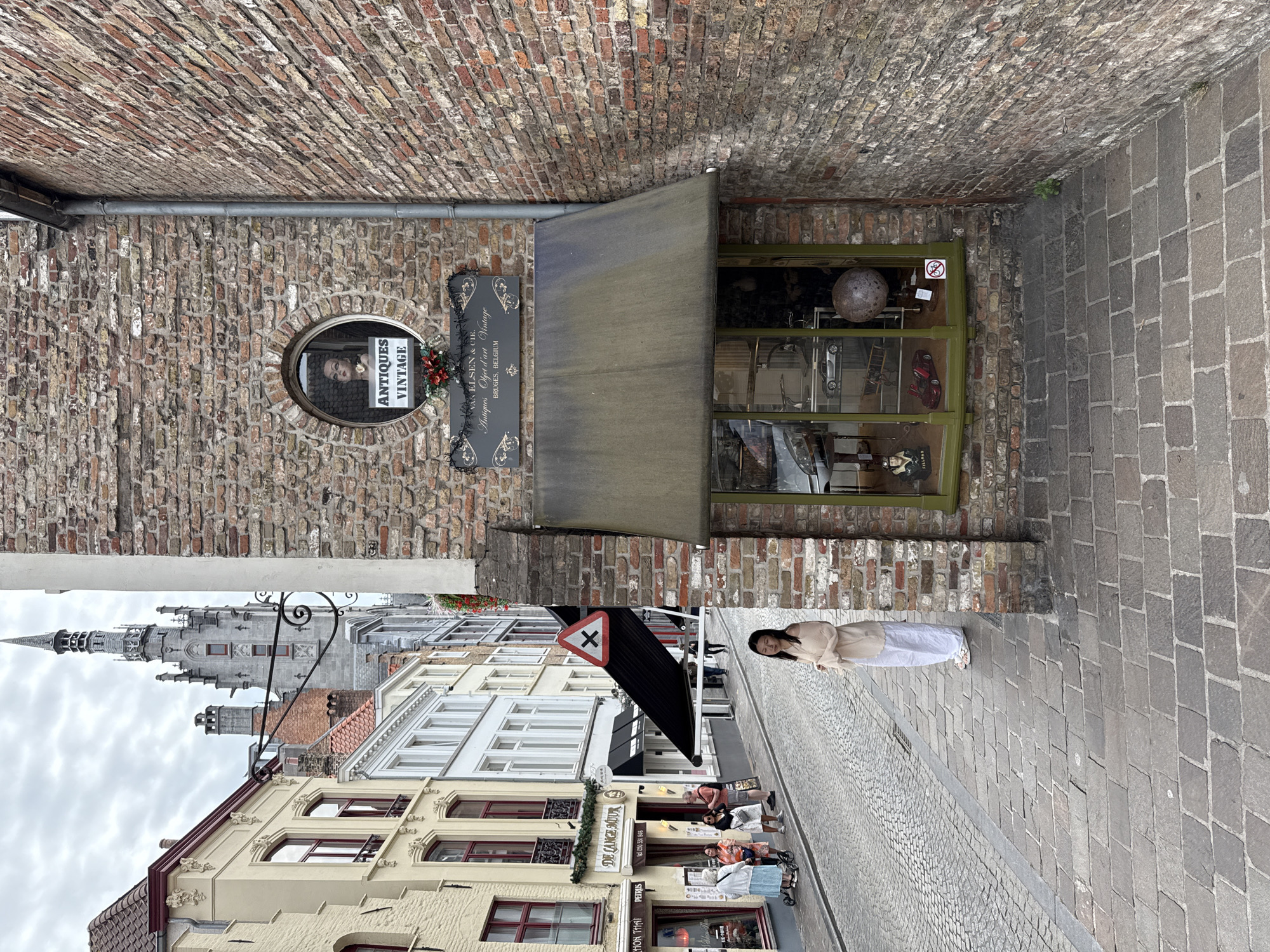 The image shows a street scene in Bruges, Belgium. On the right is the brick facade of a building with an antique shop. The shop has a green-framed window displaying various items, including a globe and model cars. Above the window, a mannequin head is visible in an oval window, with a sign below it reading "ANTIQUES VINTAGE." A dark green awning covers the shop window. On the left side of the image, a cobblestone street lined with buildings can be seen. Several people are walking on the street, and one person, a woman in a light-colored outfit, is standing on the sidewalk in the foreground. The sky is overcast.